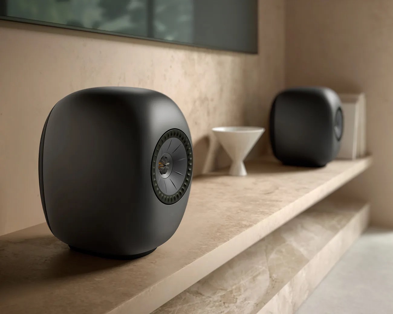 Pair of matte black rounded cube speakers on a beige marble shelf with a white decorative object between them.