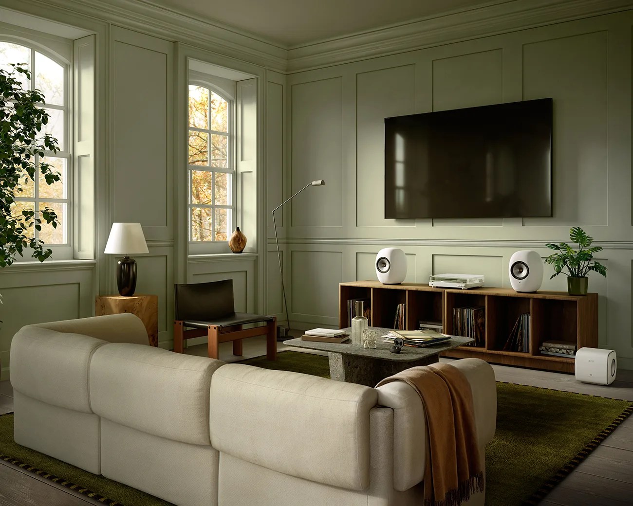 Living room with beige sofa, green rug, wooden chair, wooden media console with white speakers, and wall-mounted TV.
