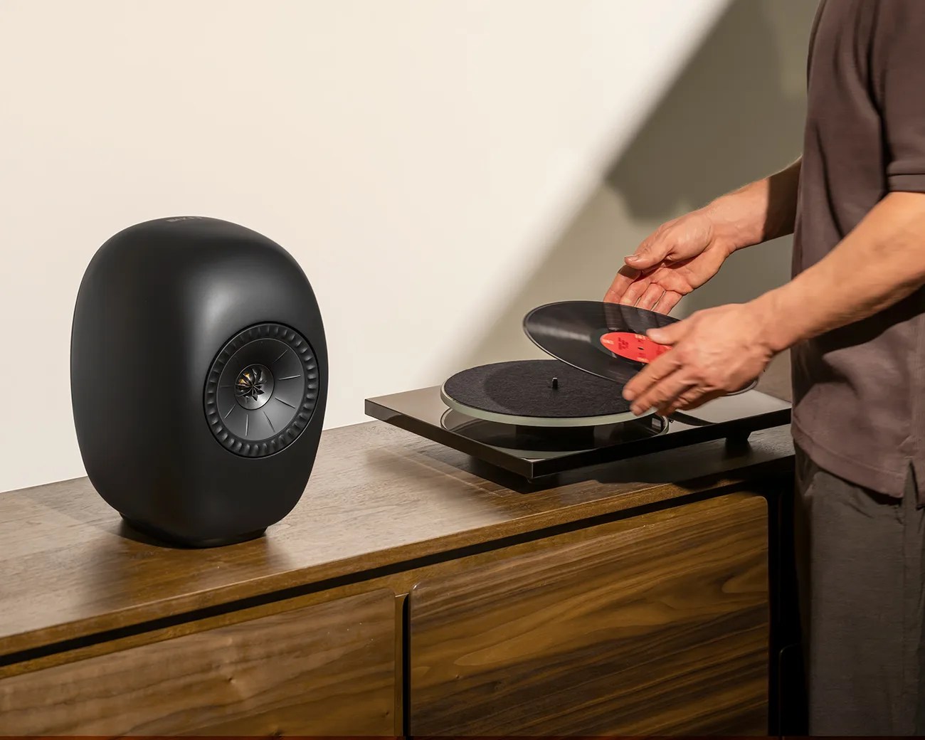 Matte black oval speaker and person placing a vinyl record on a turntable on a wooden cabinet.