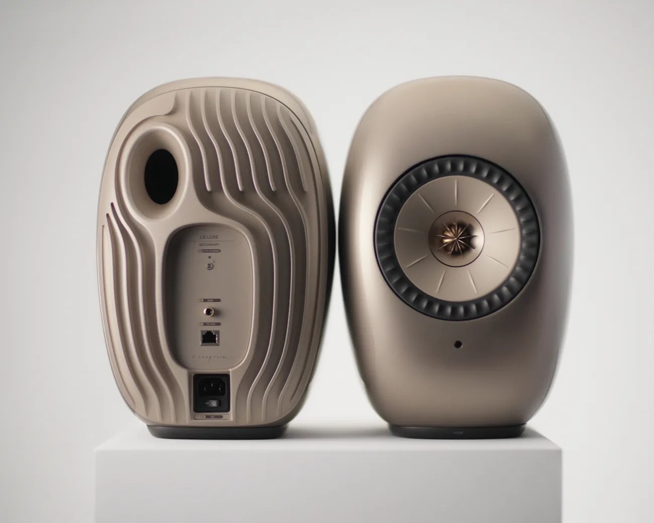 Pair of beige wireless speakers with rounded edges, one showing front speaker cone and the other showing rear ports and controls.