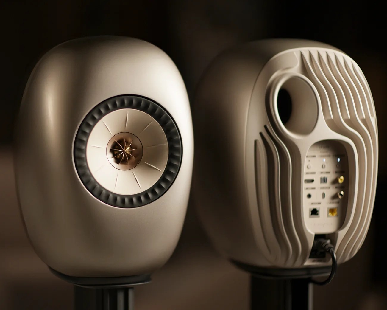 Pair of beige oval-shaped speakers with a central driver on the front and multiple input ports on the back.