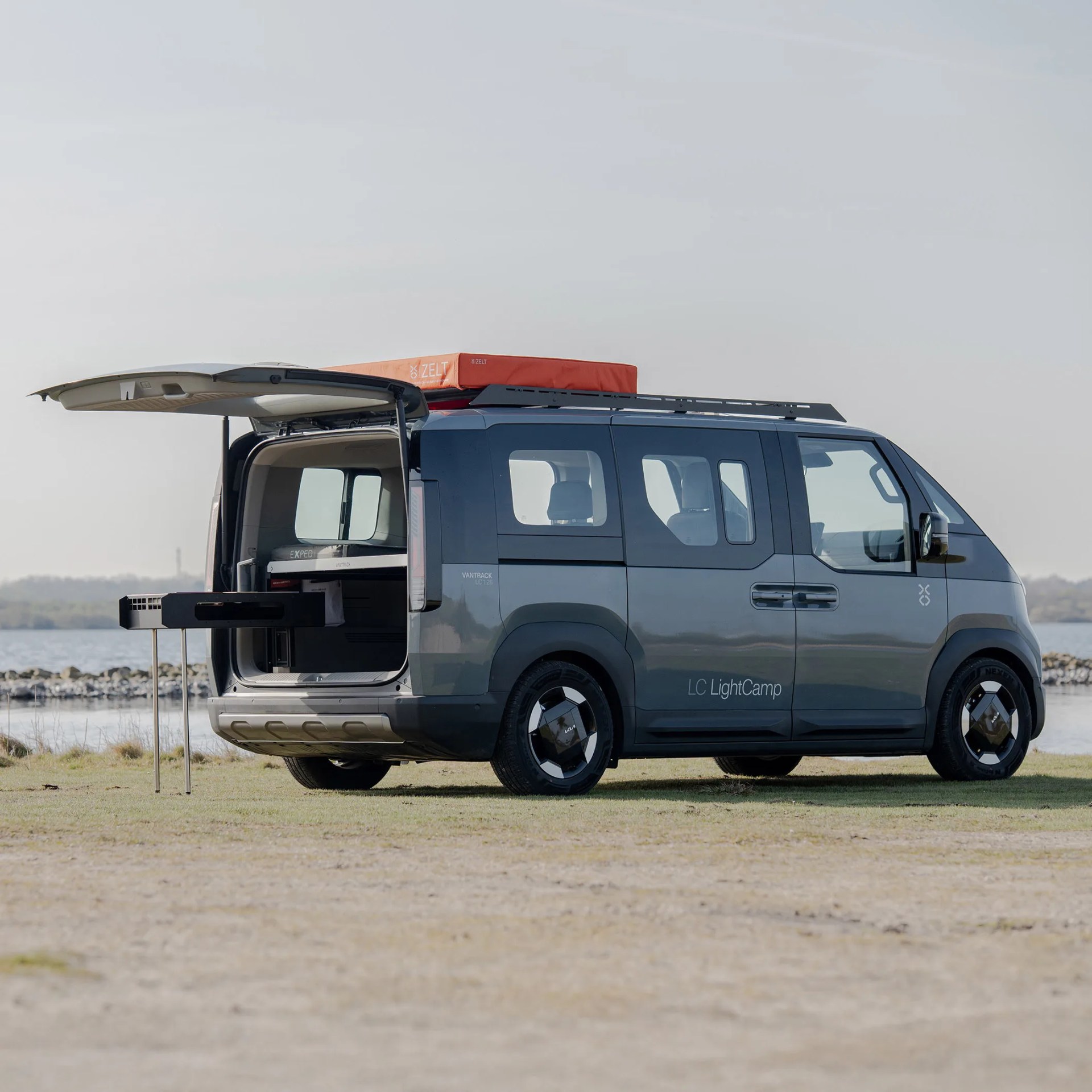 Dark gray camper van with roof tent and rear hatch open showing pull-out kitchen setup by a lakeside.