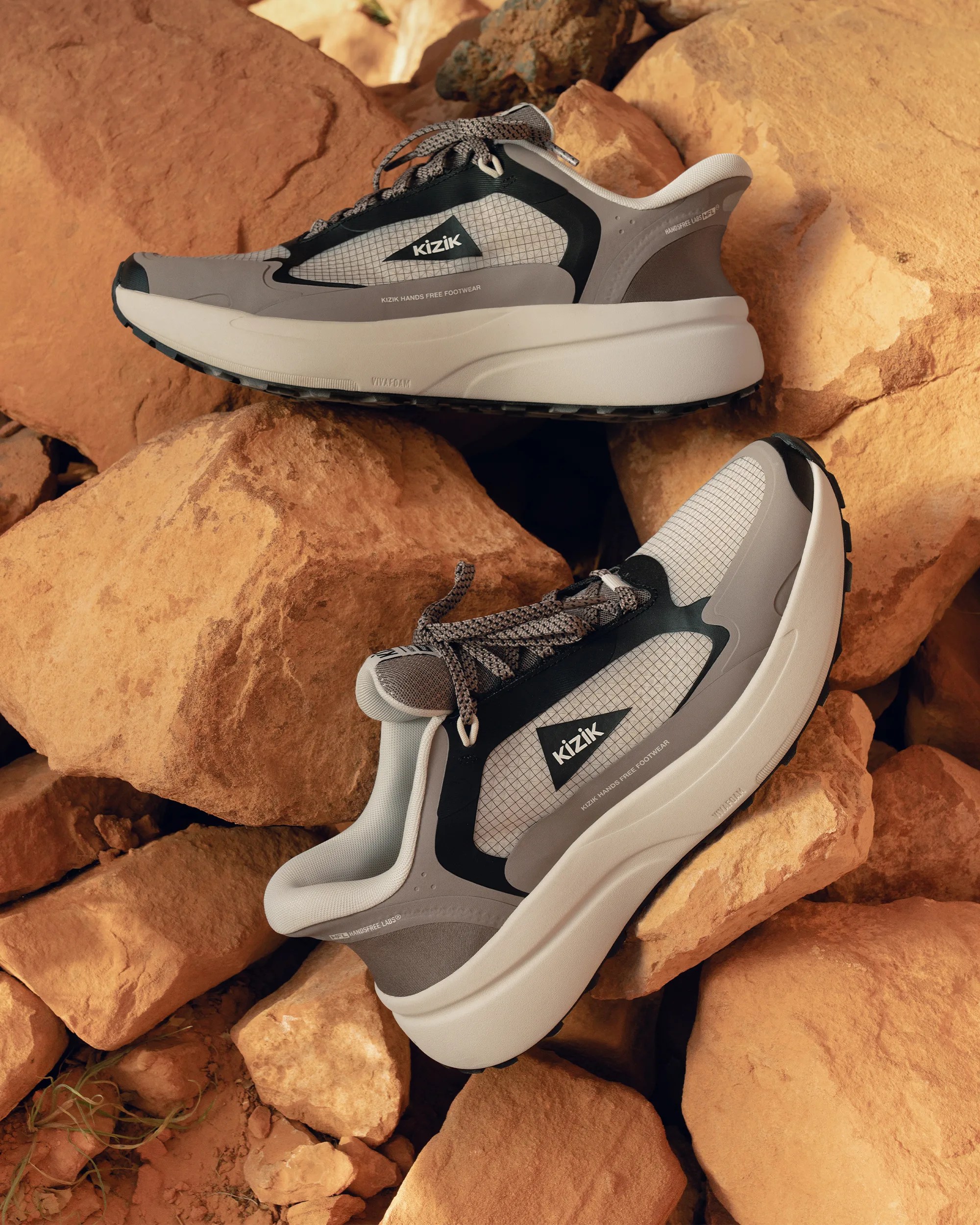 Pair of gray and black Kizik hands-free sneakers with white soles on tan rocks.
