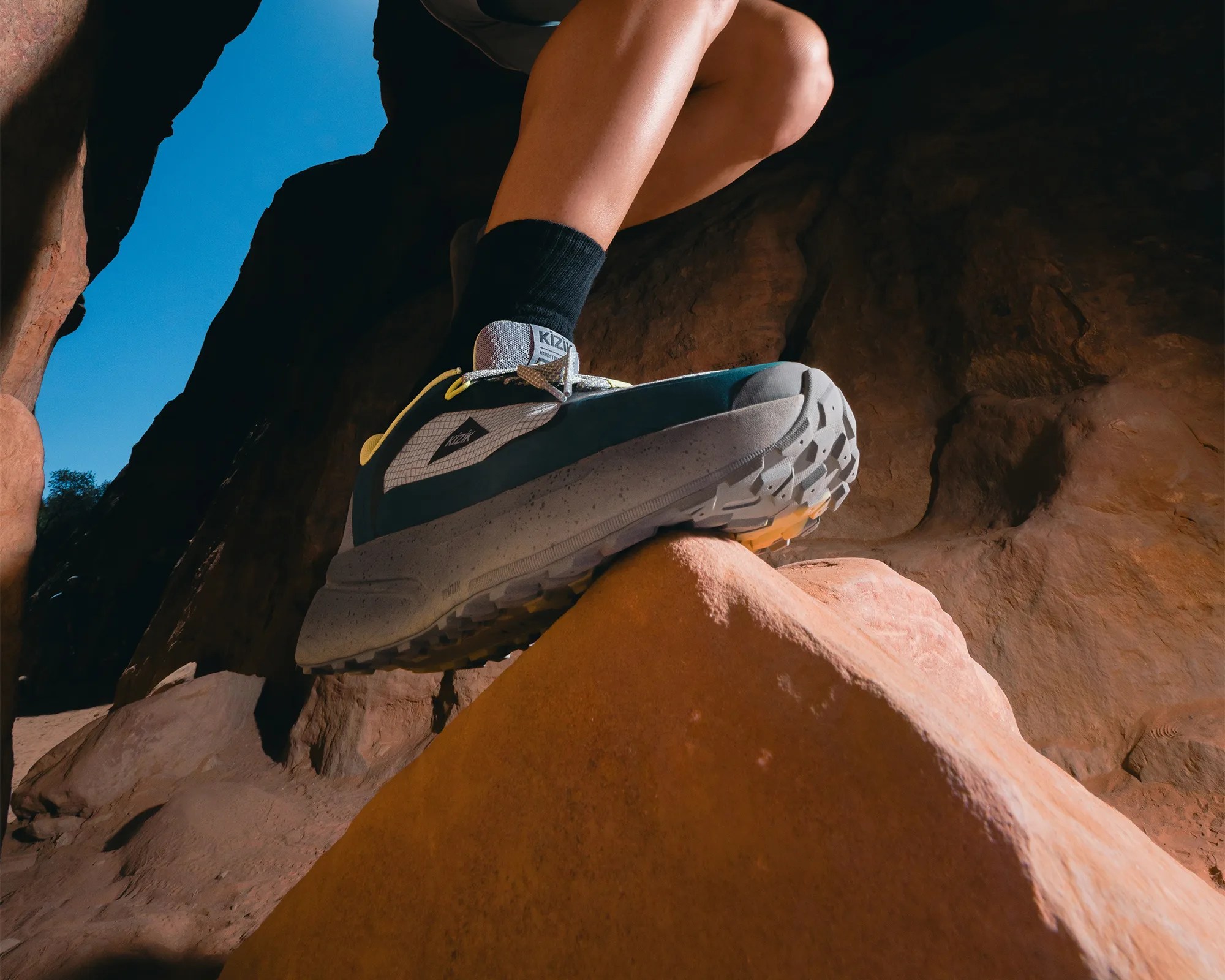 Gray and teal Kizik slip-on sneaker with black socks on a person's foot on orange rocks.