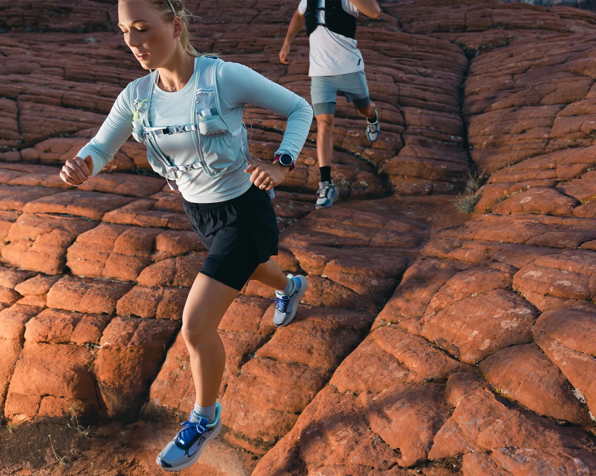 Two people trail running on reddish rocky terrain, wearing light athletic tops, hydration vests, and running shoes.