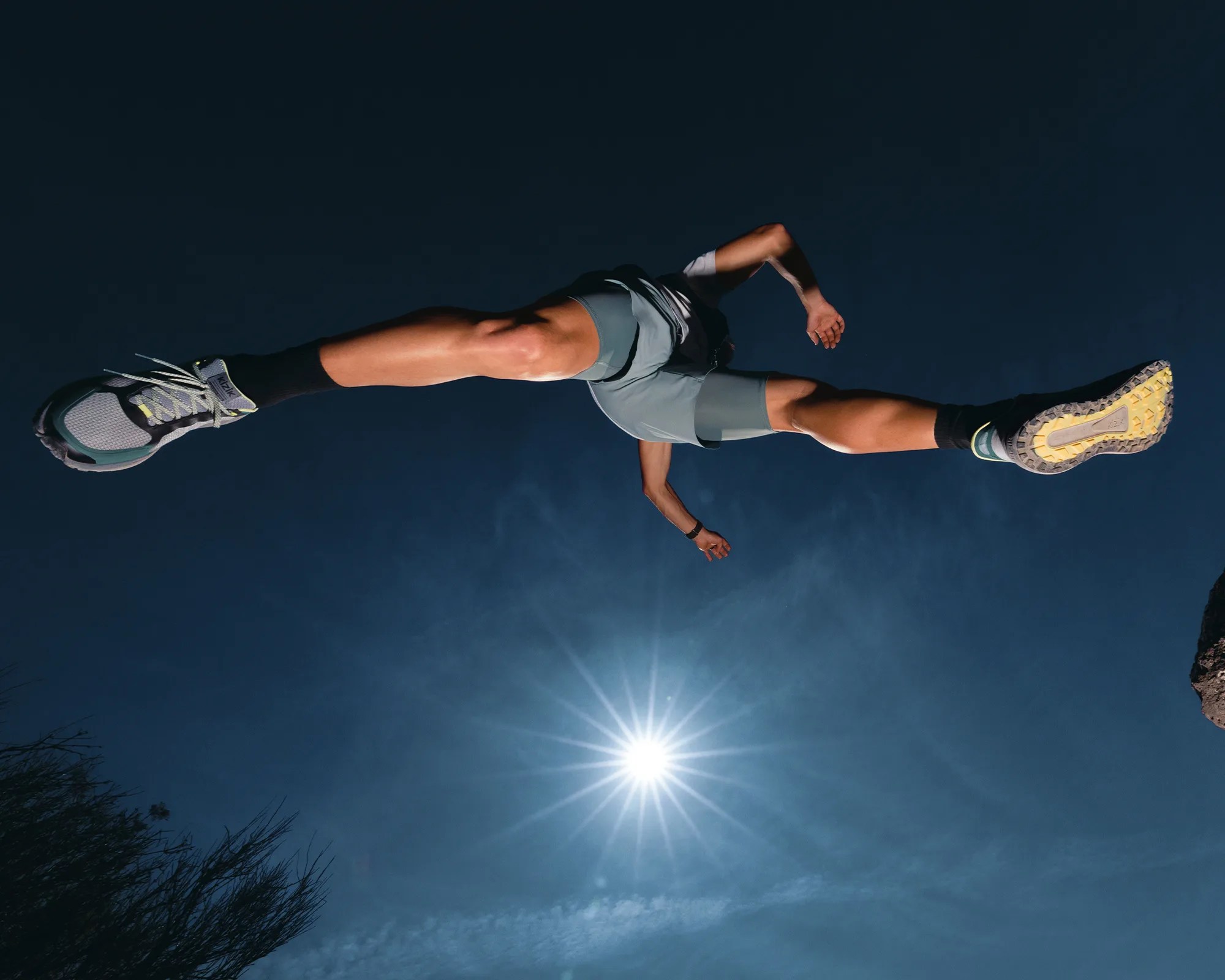 Person wearing gray shorts and black socks with gray and yellow running shoes jumping against a clear blue sky with the sun visible.