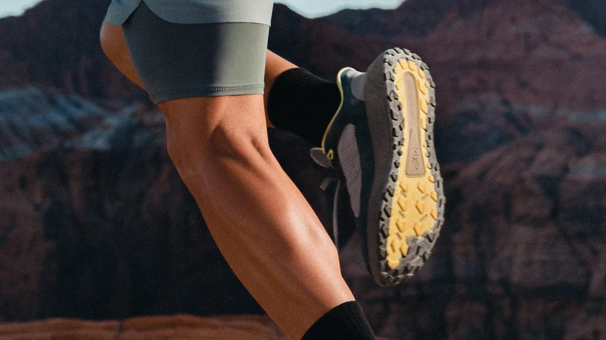 Runner's leg in gray shorts and black socks wearing black and yellow trail running shoes with rugged soles.