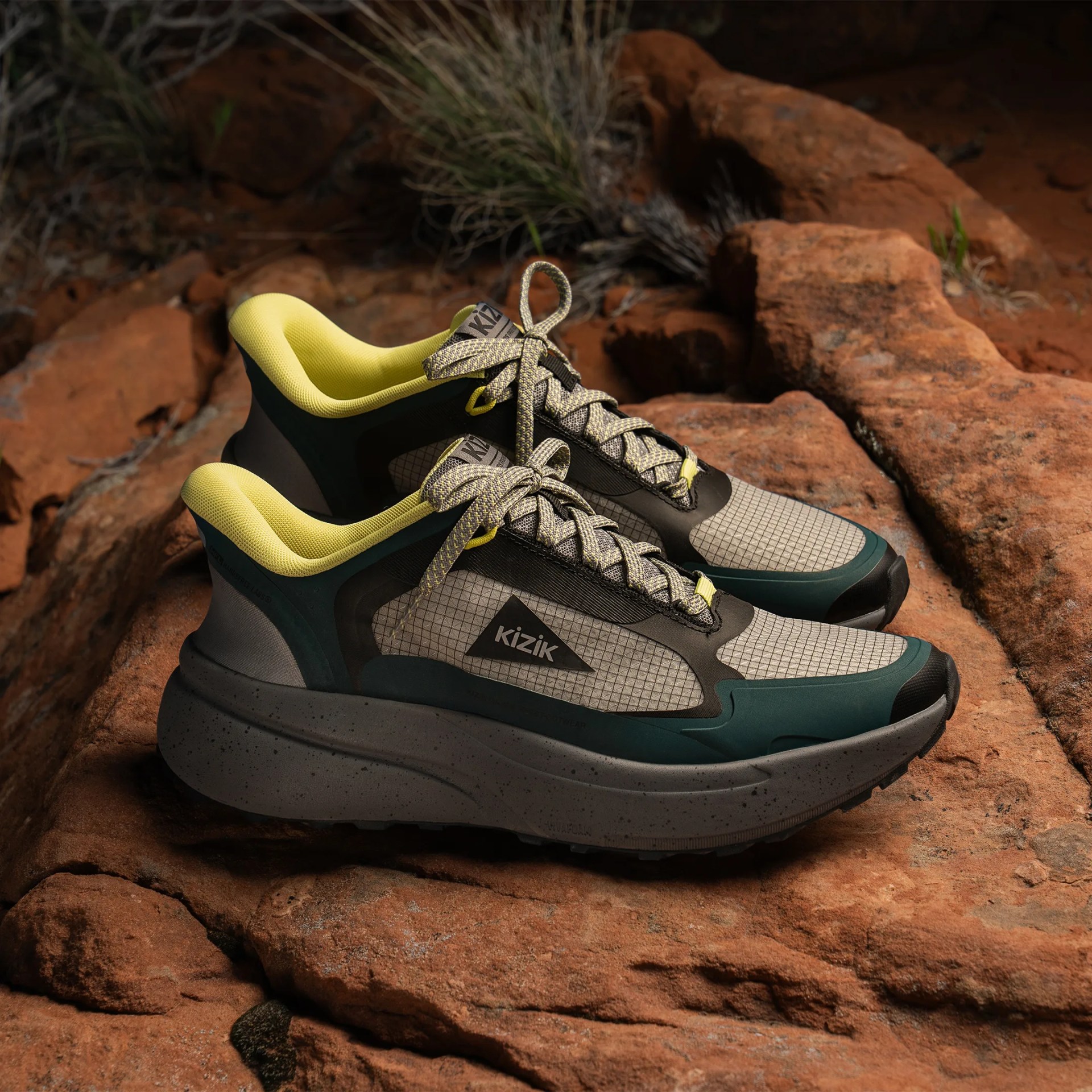 Pair of Kizik brand sneakers with yellow interior lining and gray-green exterior on rocky ground.