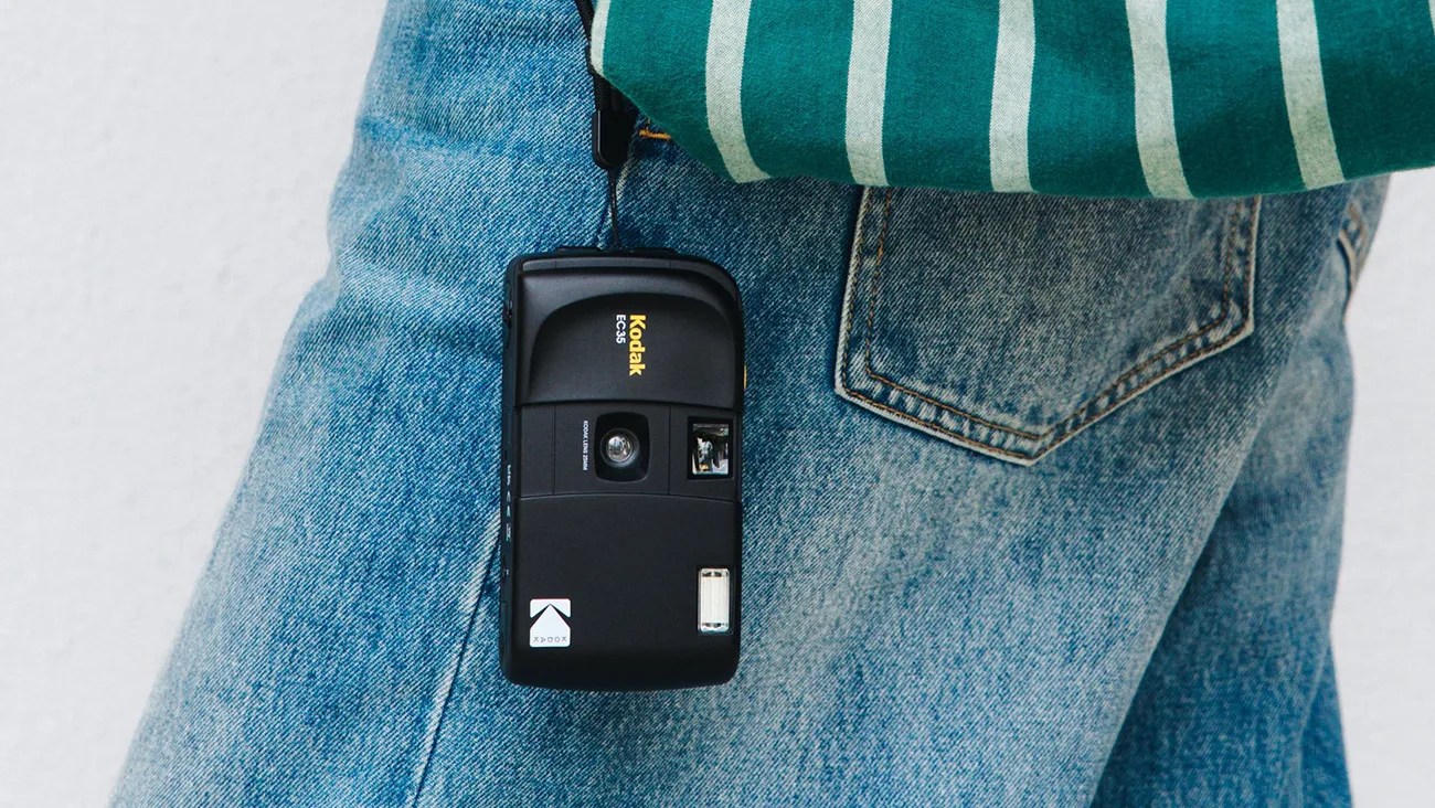 Black Kodak EC35 film camera hanging from a wrist strap against blue denim jeans.