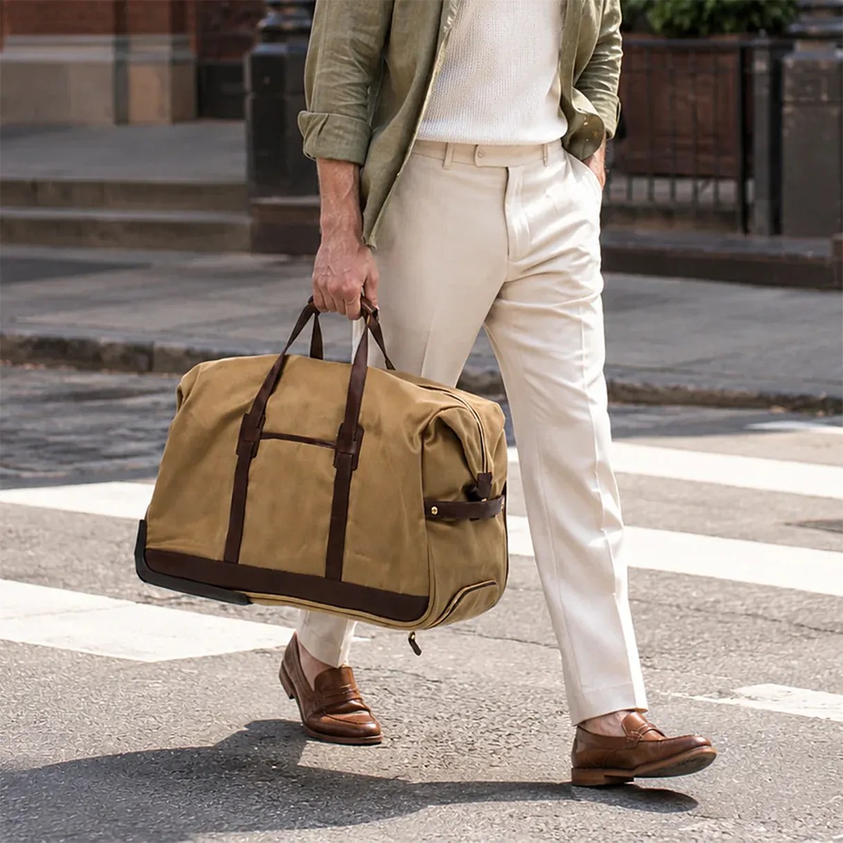 Tan canvas duffel bag with dark brown leather handles and trim held by a person wearing cream pants and brown leather loafers.