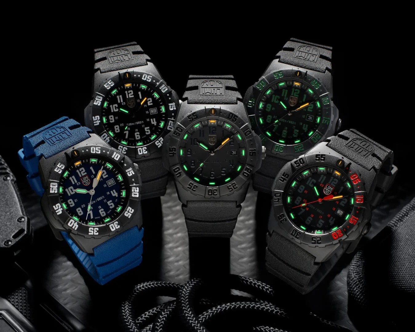 Five Luminox wristwatches with black cases and illuminated dials, four with black straps and one with a blue strap, arranged on a dark surface.