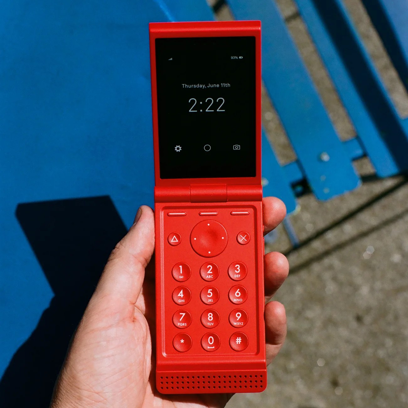 Hand holding a red flip phone with a digital screen showing the time 2:22 and a numeric keypad.