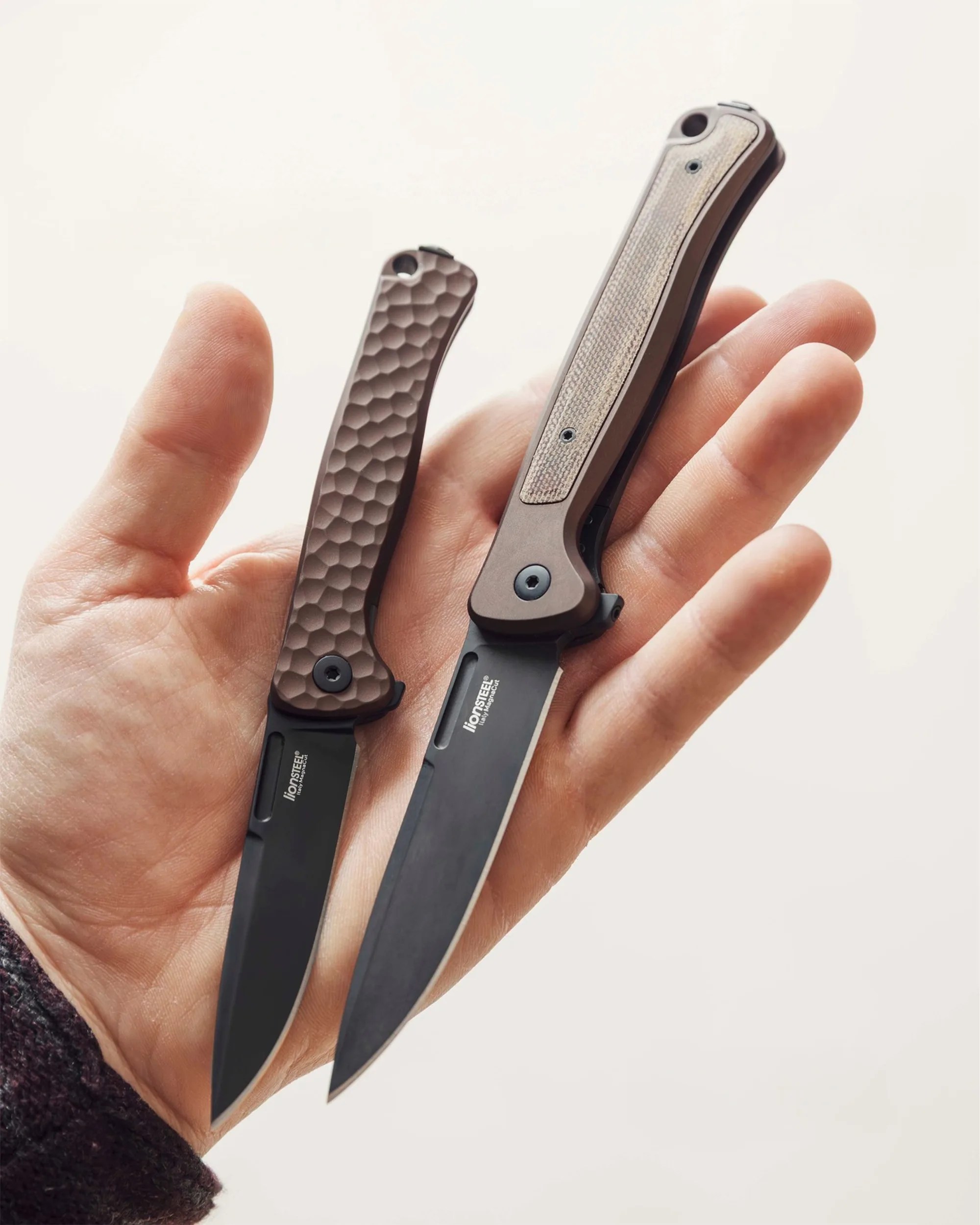 Hand holding two folding knives with black blades and textured brown handles.