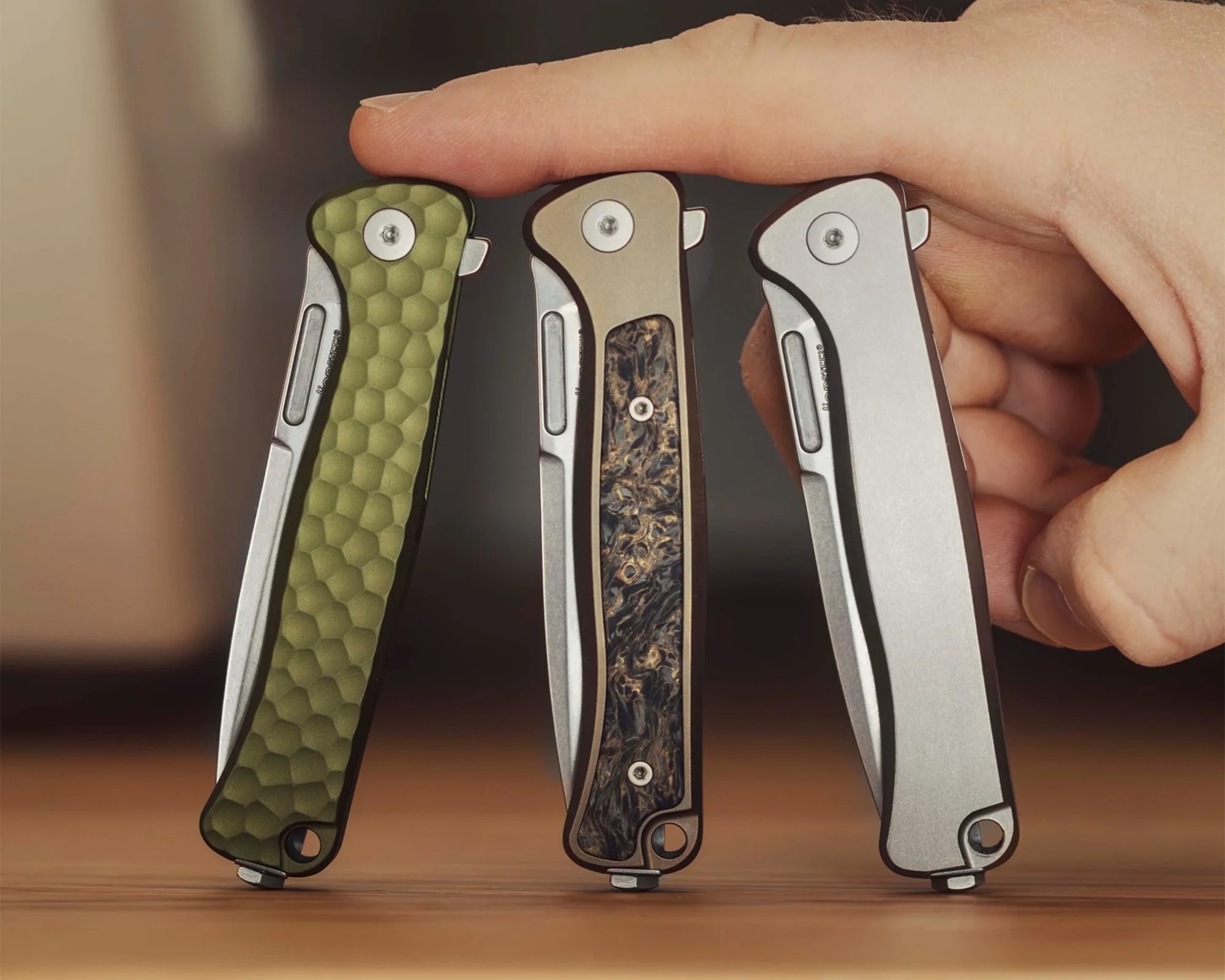Three folding knives with different handle textures and colors—green honeycomb, brown marbled, and plain silver—held upright on a wooden surface.