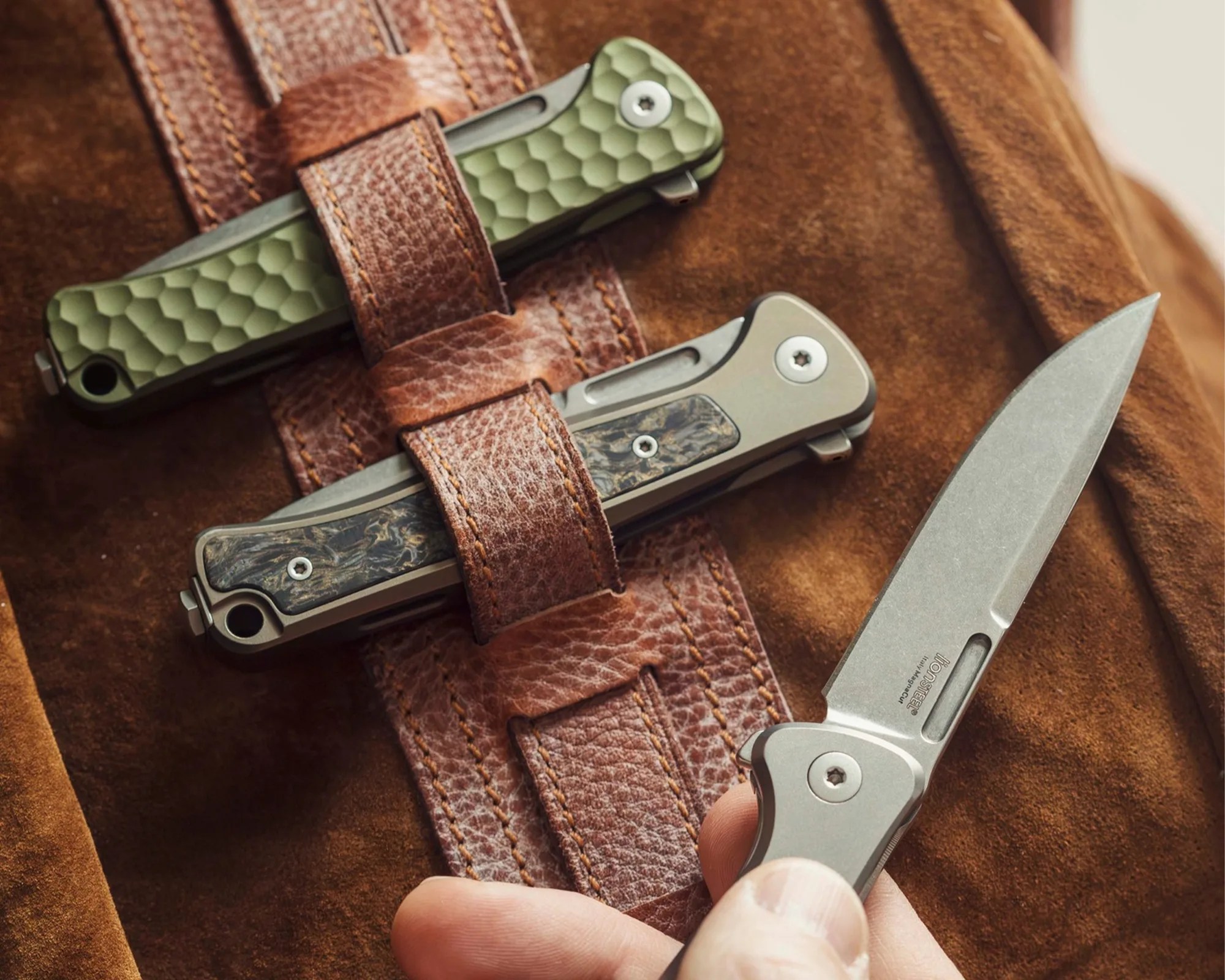 Three folding knives with textured handles secured by brown leather straps on a suede surface, one knife blade partially open in a hand.