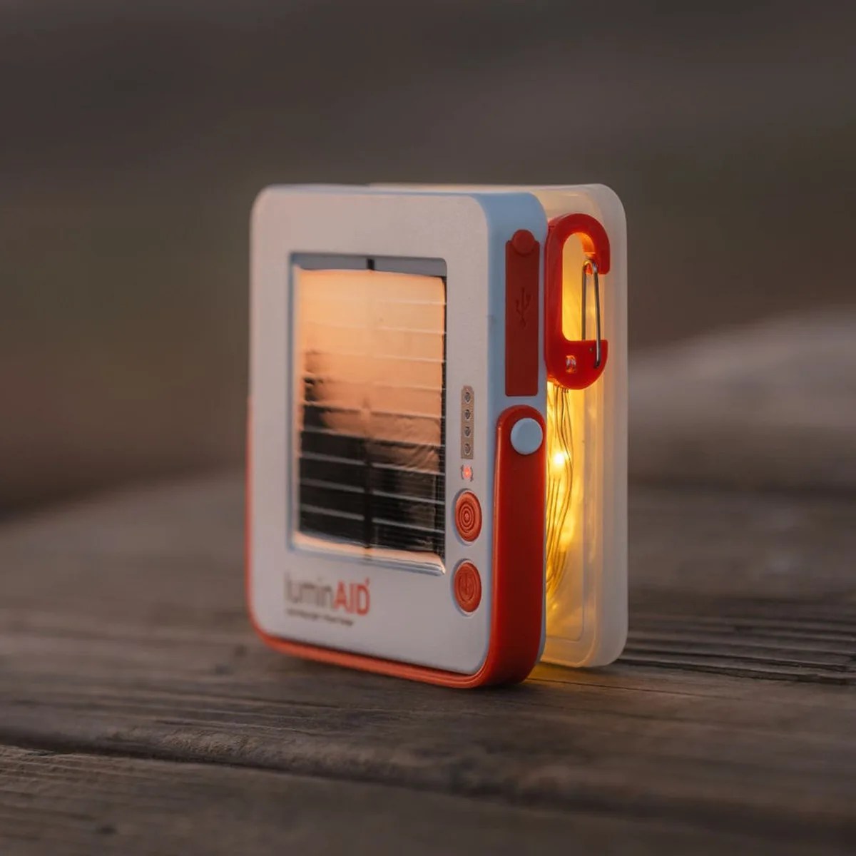 Compact white and orange solar-powered lantern with a glowing light and a red carabiner clip on a wooden surface.