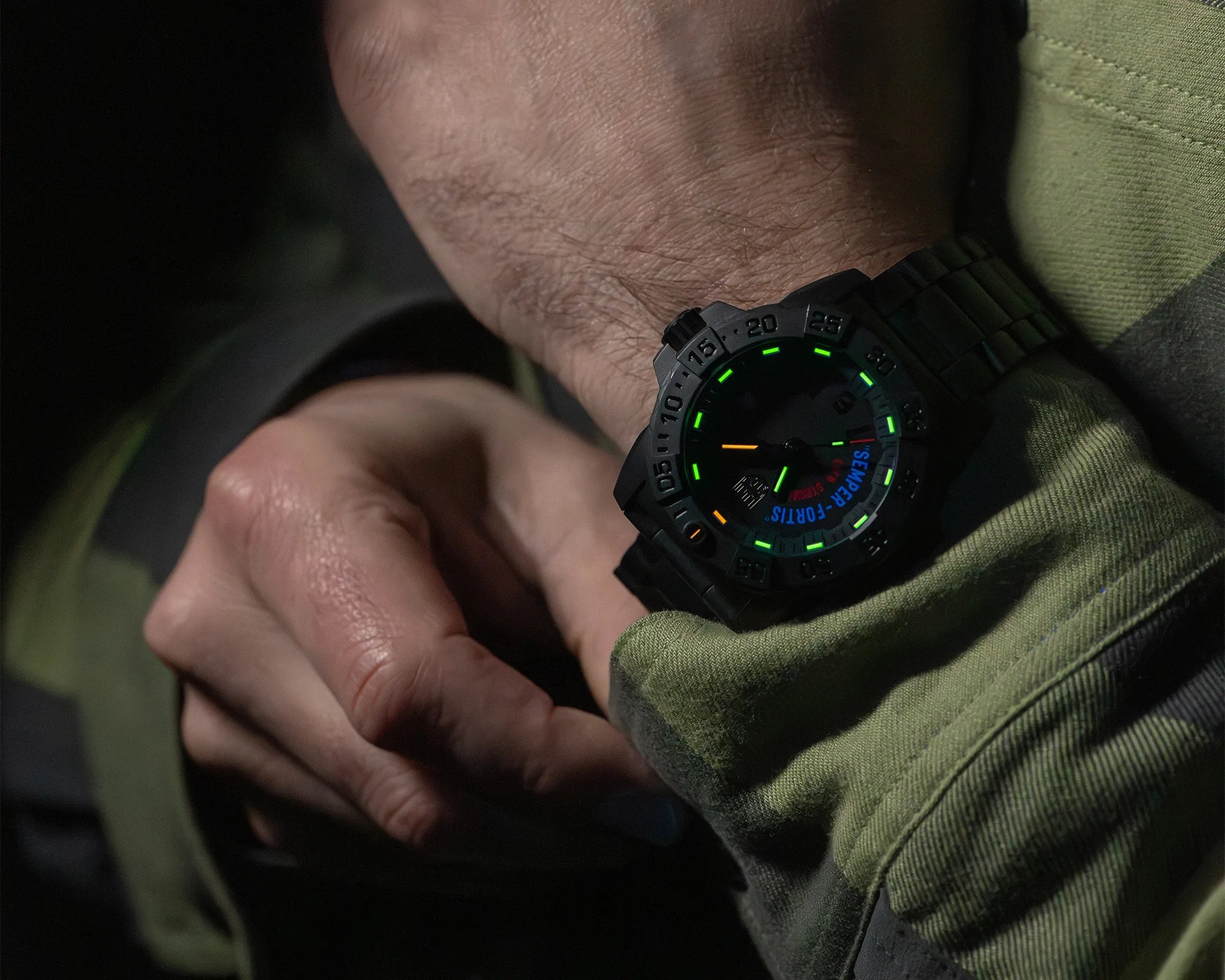 Black wristwatch with glowing green and orange markers worn on a wrist over a green fabric sleeve.