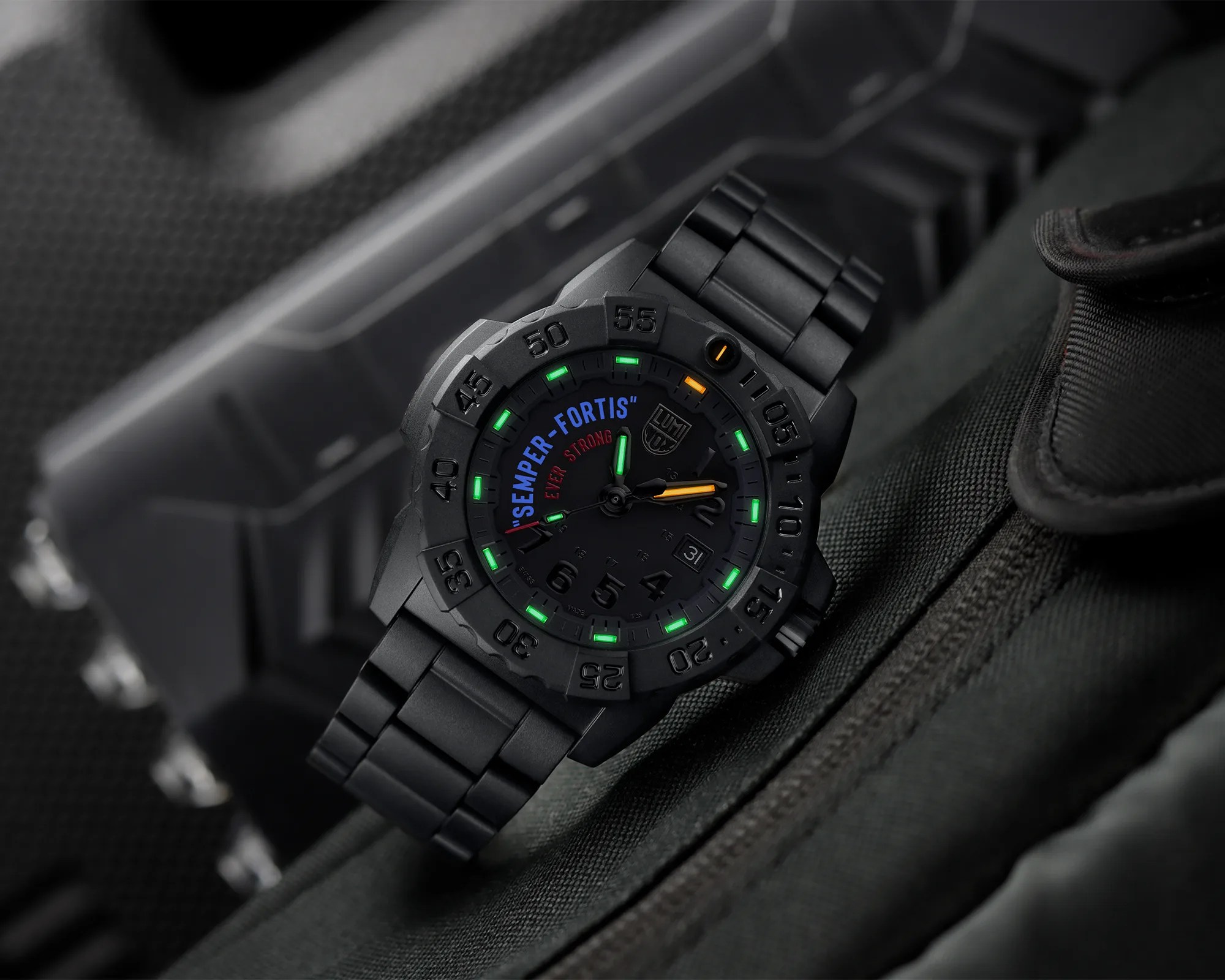 Black tactical wristwatch with luminous green and orange markers and a metal link band on dark fabric.