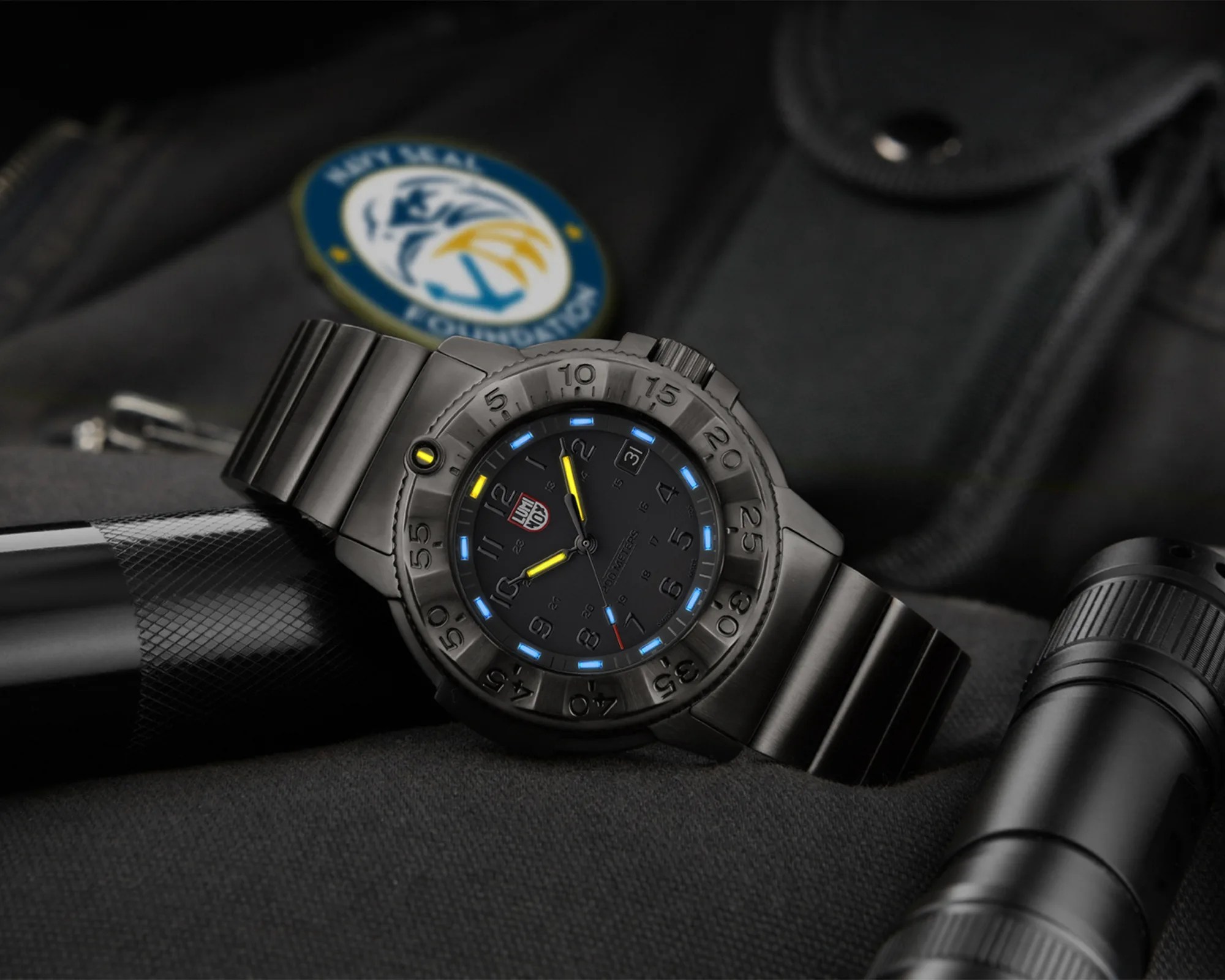Black tactical wristwatch with blue and yellow luminescent markers on a black fabric surface near a Navy SEALs patch.