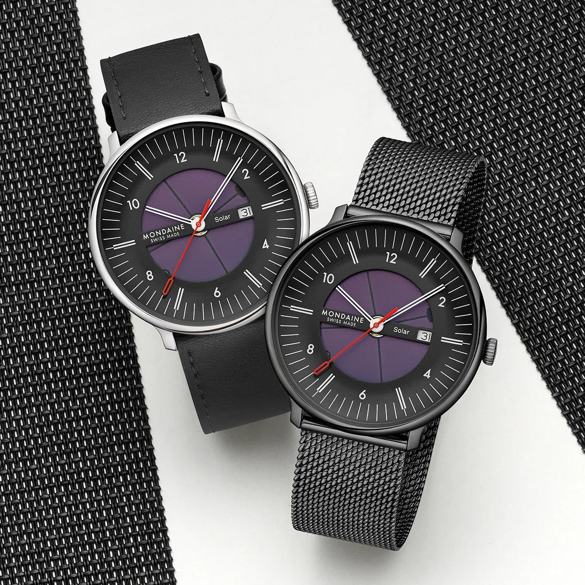 Two Mondaine Swiss Made solar watches with black and silver cases, purple dials, and black leather and mesh straps on a white surface.