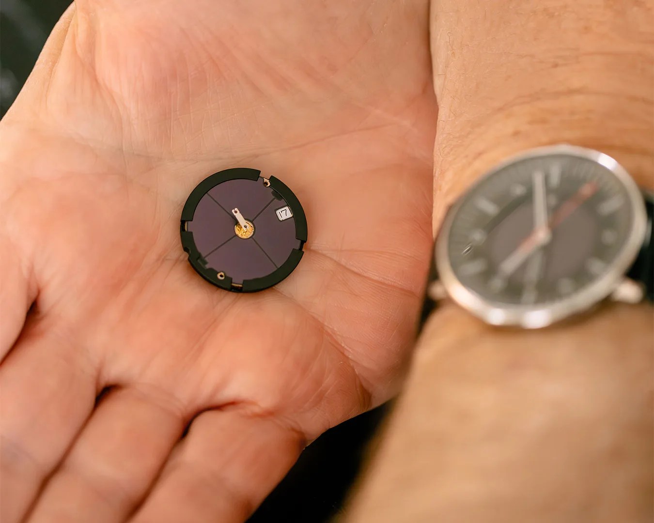 Small black watch face with white hands and date display held in a palm next to a wristwatch on a wrist.