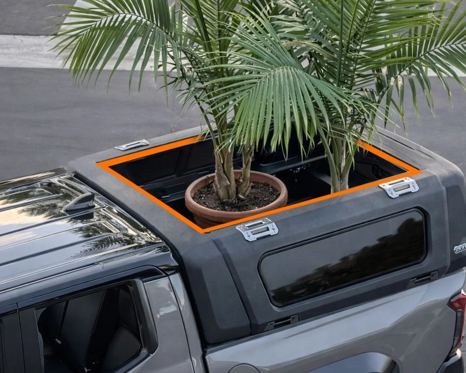 Gray pickup truck with a black camper shell featuring a large rectangular sunroof opening with an orange trim, showing a potted palm plant inside.