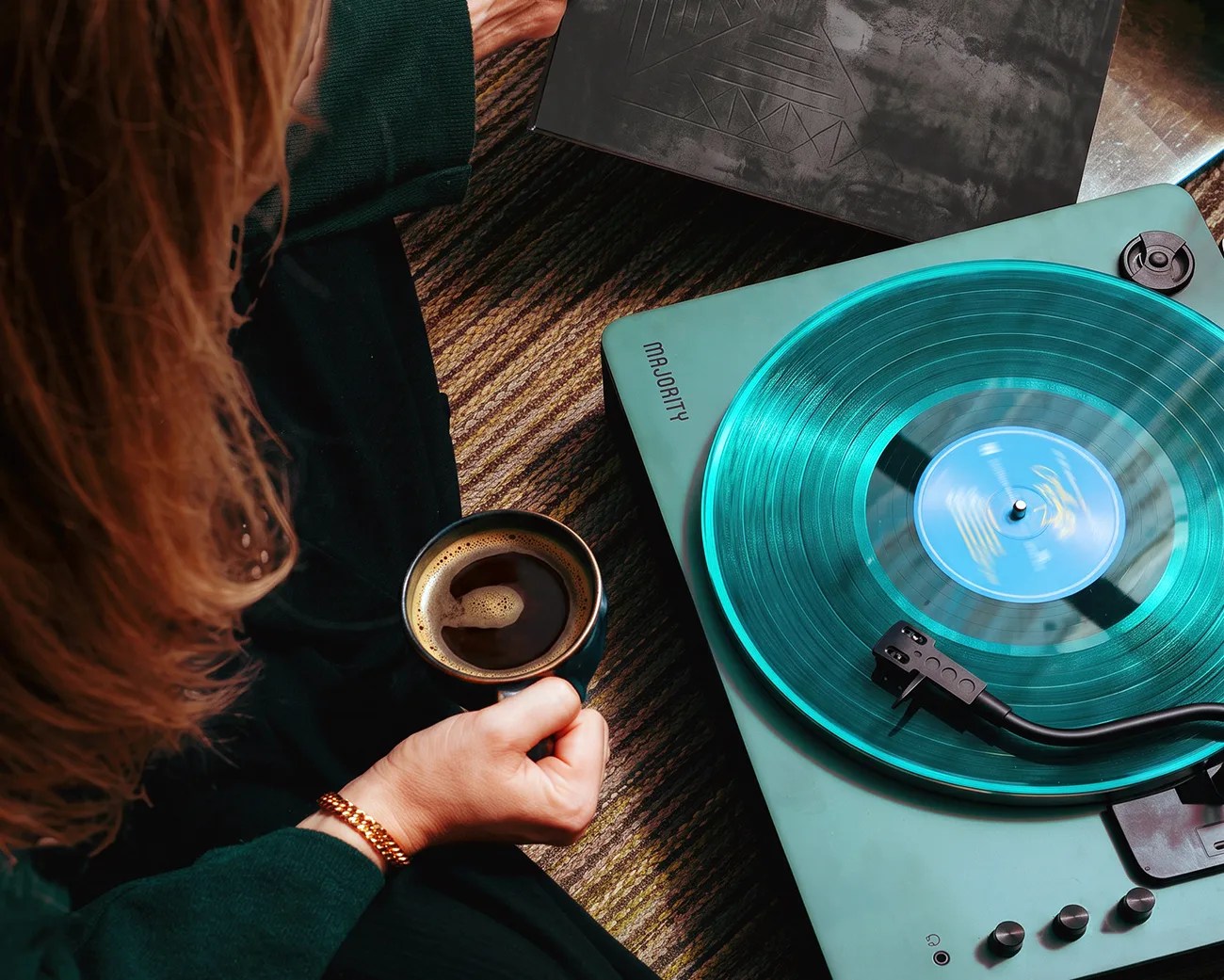 Person holding a cup of coffee next to a teal Majority turntable playing a translucent blue vinyl record.
