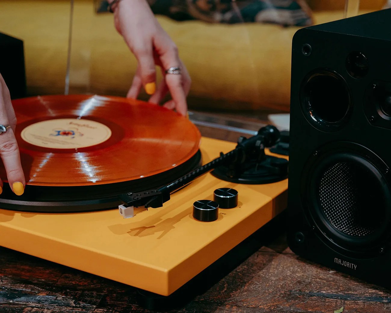 Yellow turntable with an orange vinyl record being placed on it, next to a black Majority speaker on a wooden surface.