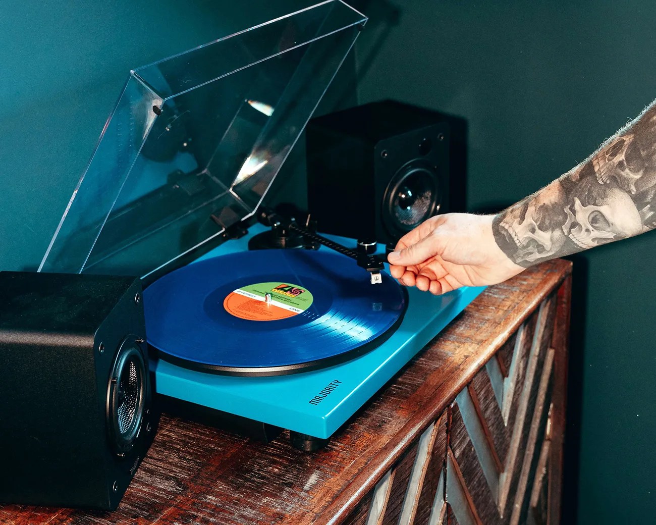 Blue Majority turntable with a blue vinyl record playing, flanked by black speakers on a wooden cabinet, with a tattooed arm adjusting the tonearm.