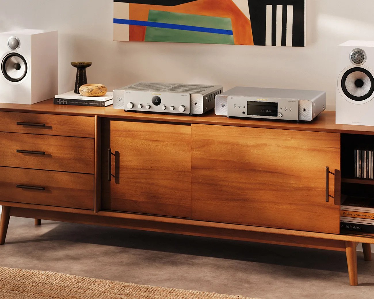 Mid-century wooden sideboard with silver audio amplifier, CD player, white speakers, books, and decorative items on top.