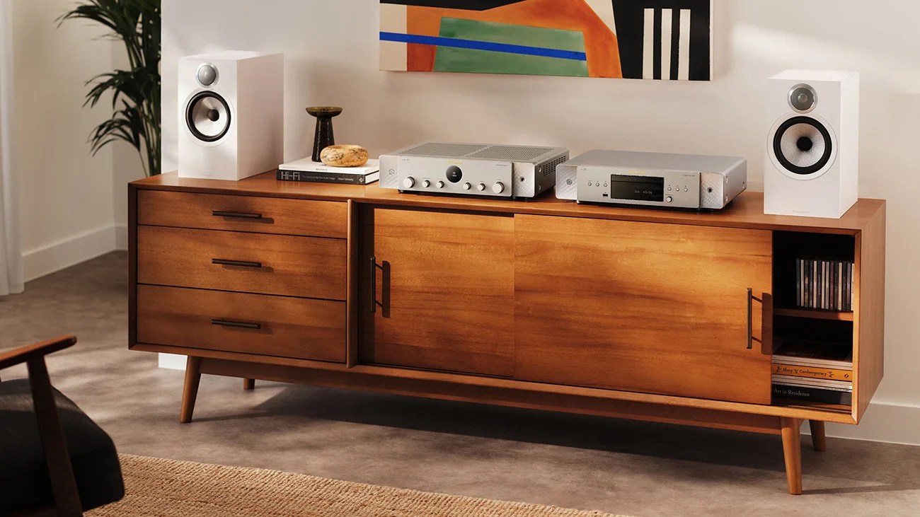 Mid-century modern wooden sideboard with three drawers, two sliding doors, white bookshelf speakers, and silver audio equipment on top.