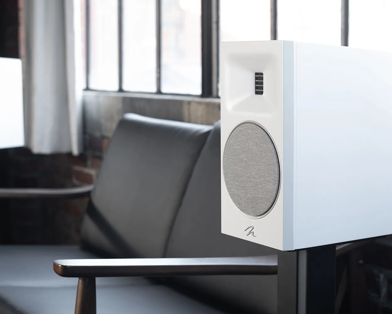 White bookshelf speaker with gray fabric woofer grille on a black stand in front of a black leather sofa.