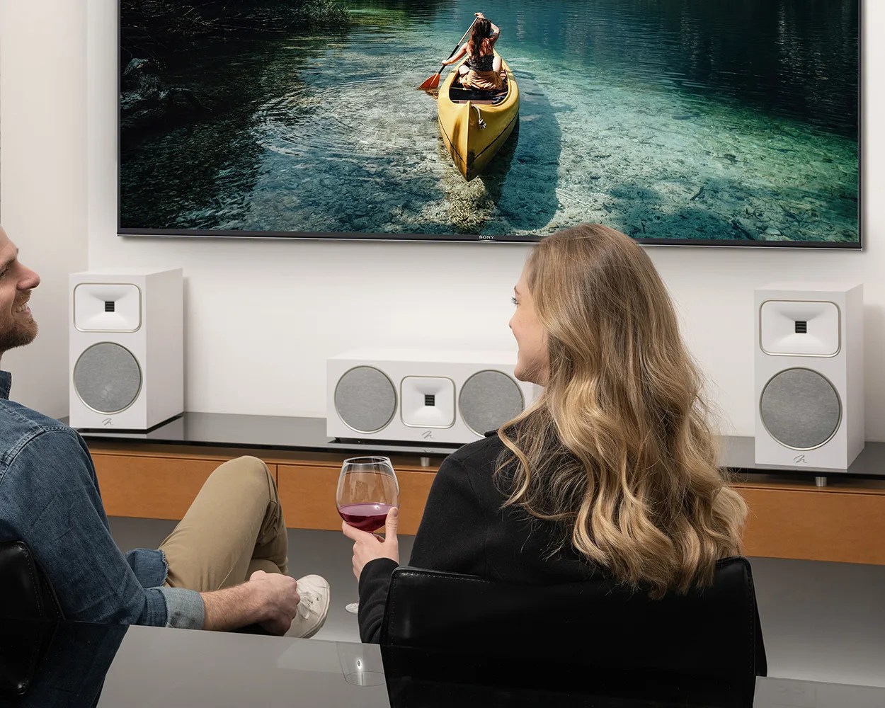 Two people sitting in black chairs watching a wall-mounted Sony TV with a yellow kayak on clear water, with white speakers on a wooden cabinet below.