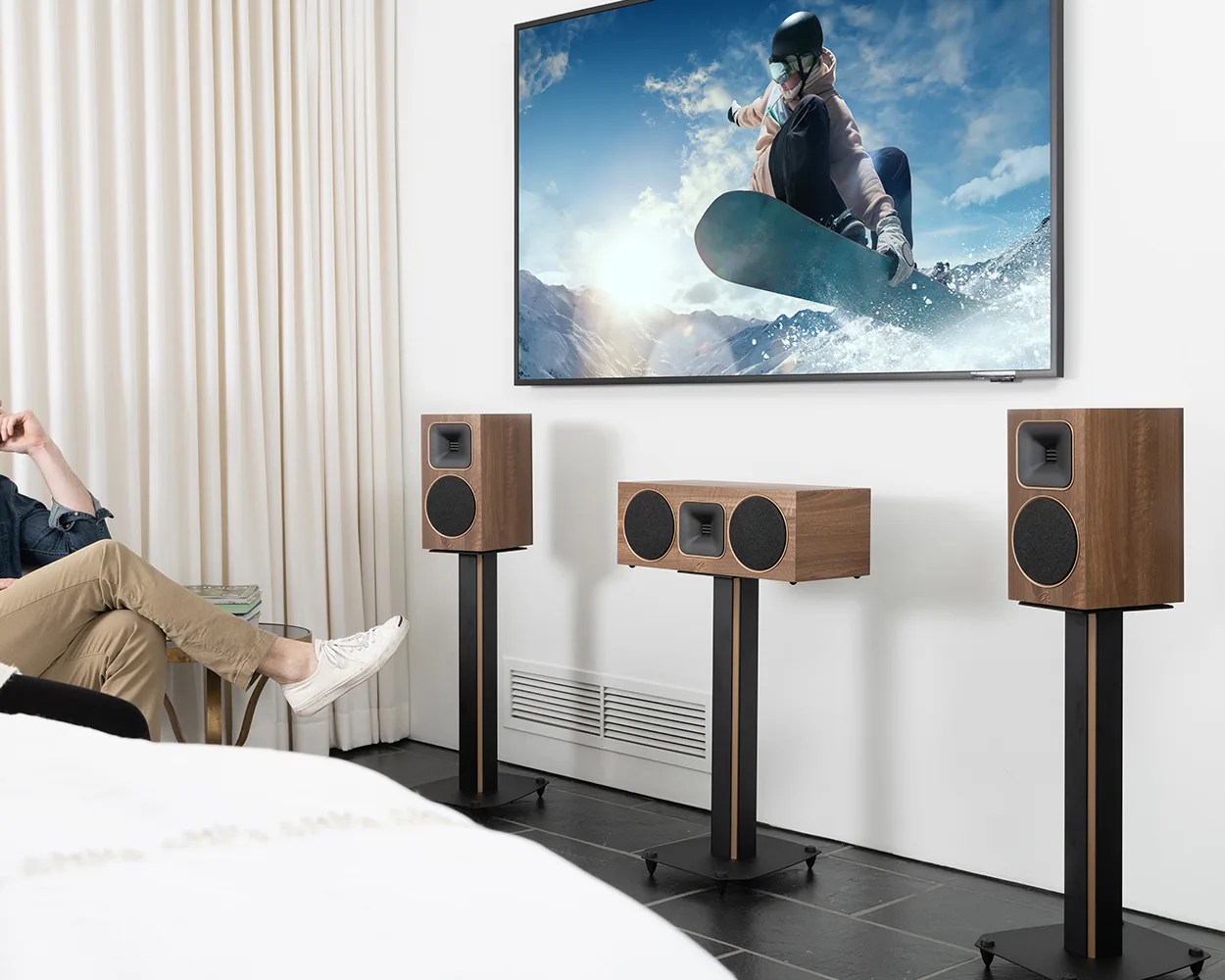 Three wooden speakers on black stands arranged below a wall-mounted TV showing a snowboarder in action.