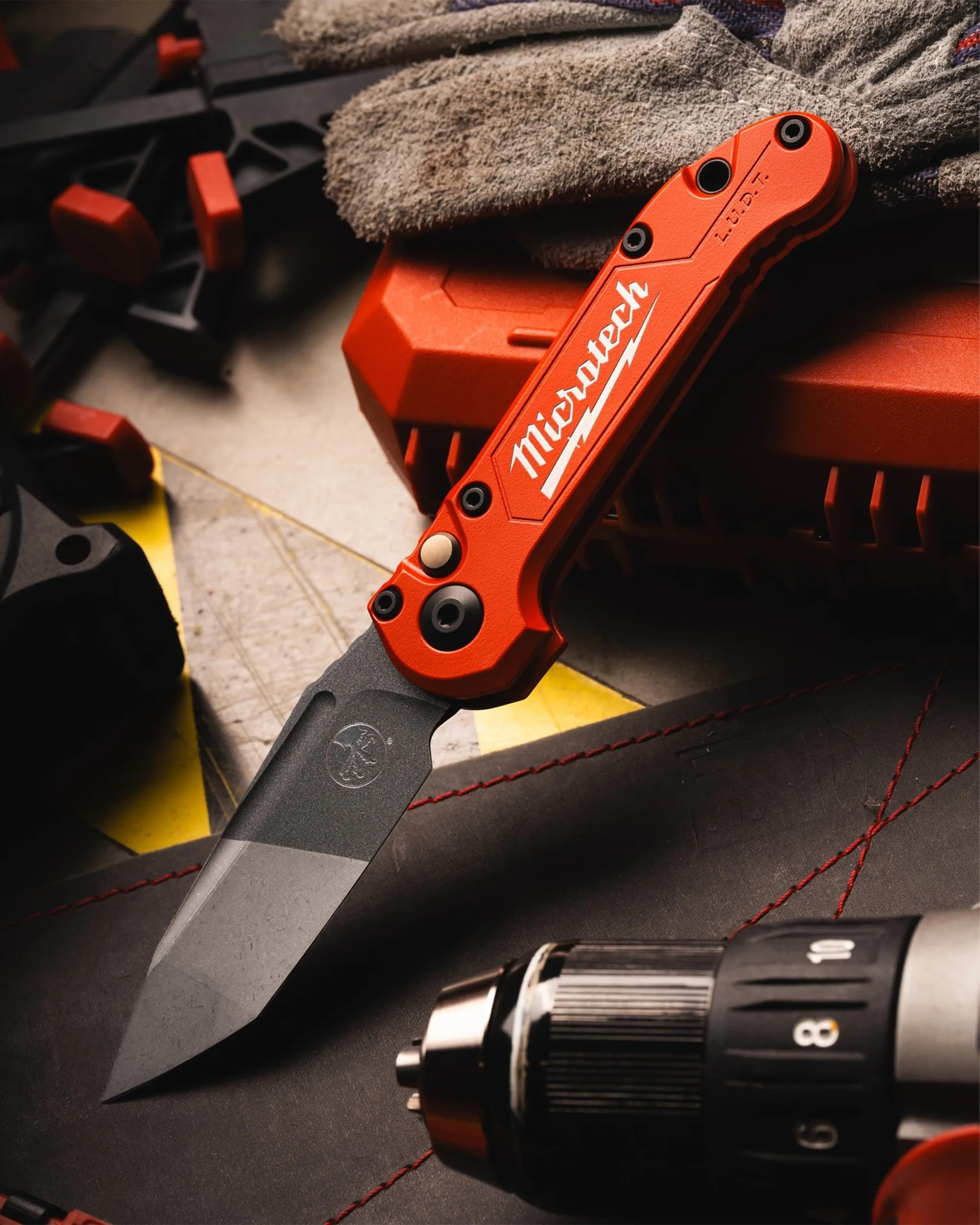 Orange-handled Microtech folding knife with a black blade resting on a workbench near gloves and a power tool.