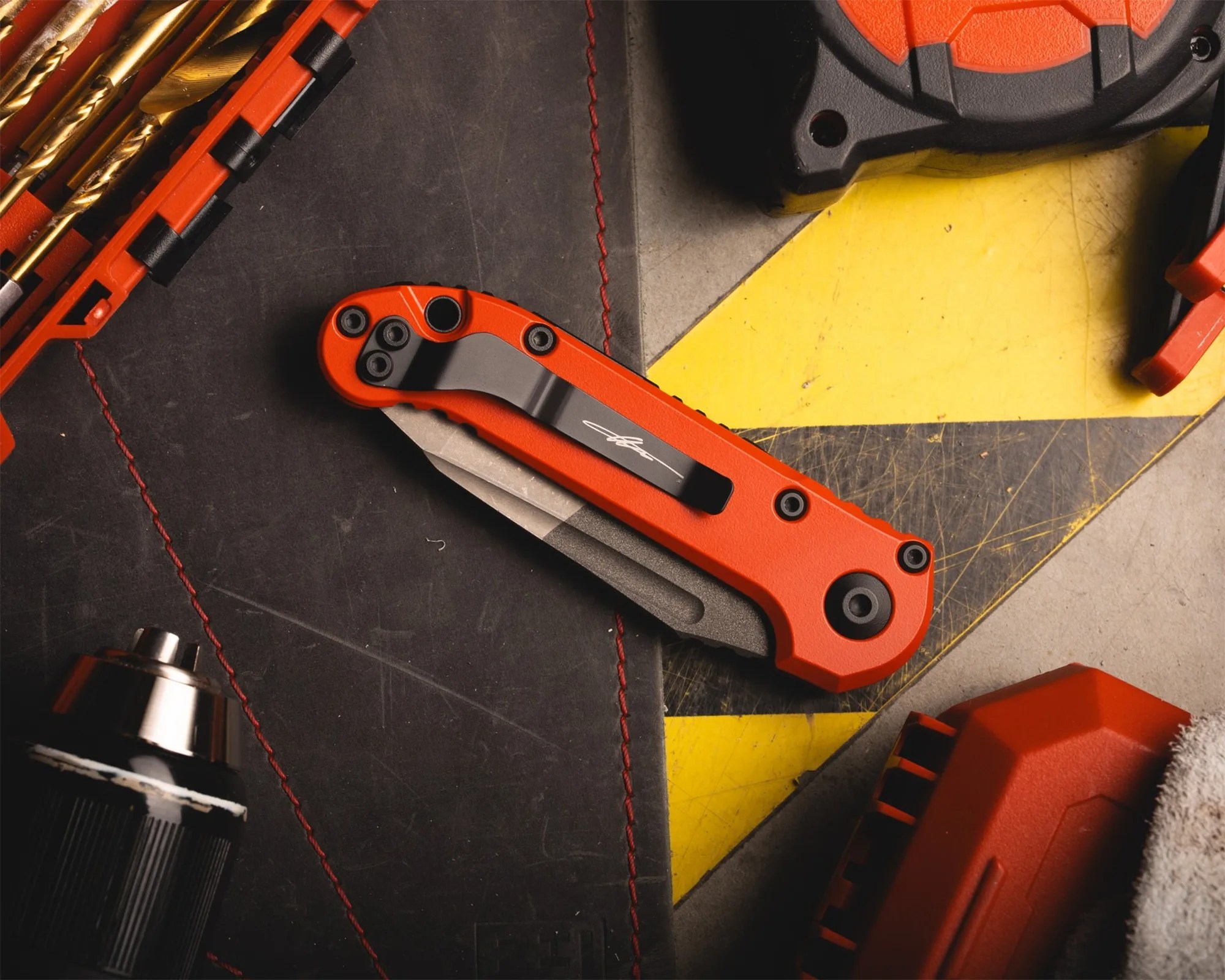 Orange folding knife with black clip on a black and yellow work surface next to drill bits and a power tool.