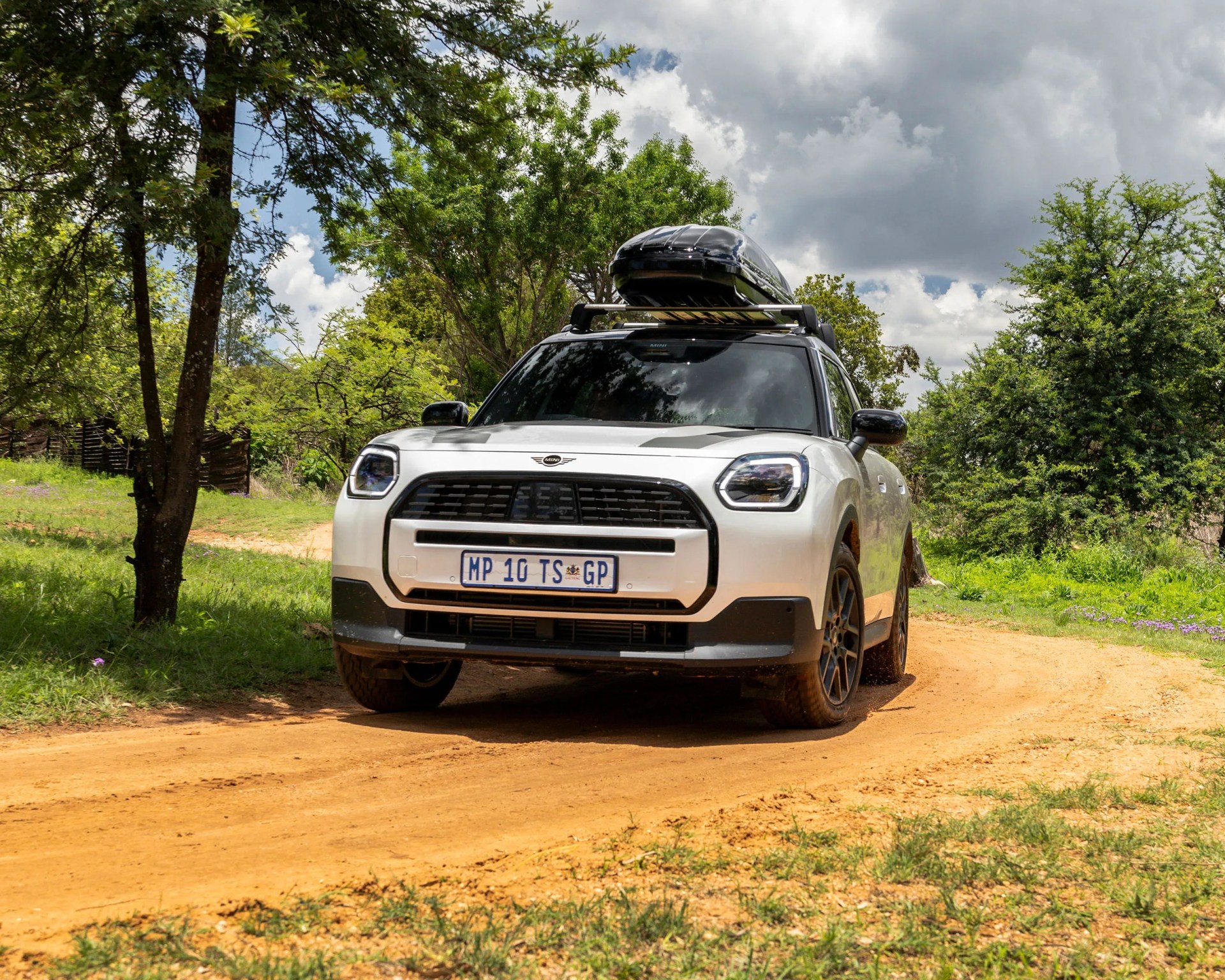 White Mini SUV with black roof box on a dirt road surrounded by green trees under a cloudy sky.