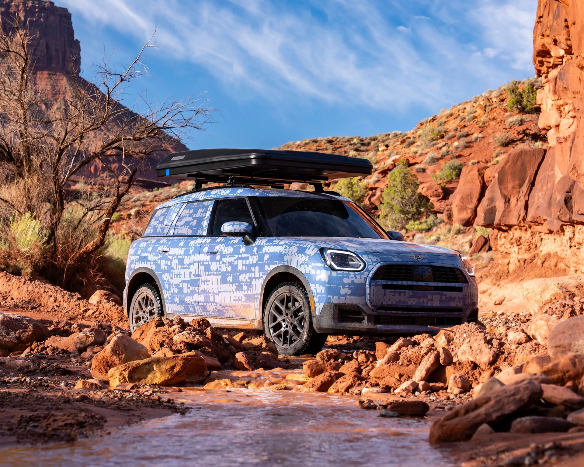Blue and white camouflaged SUV with black roof cargo box driving on rocky terrain near a stream.