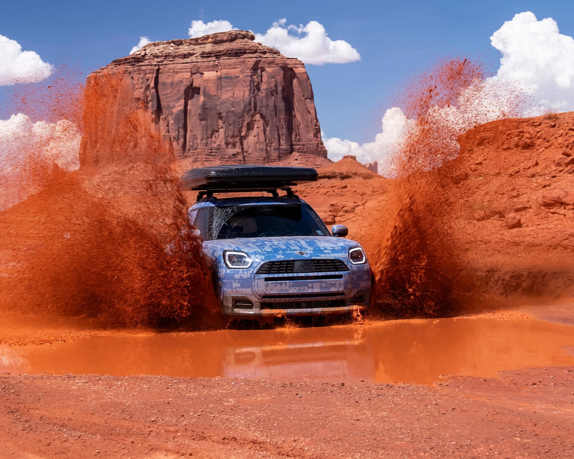 Blue camouflaged SUV splashing red mud in a desert landscape with large rock formations.