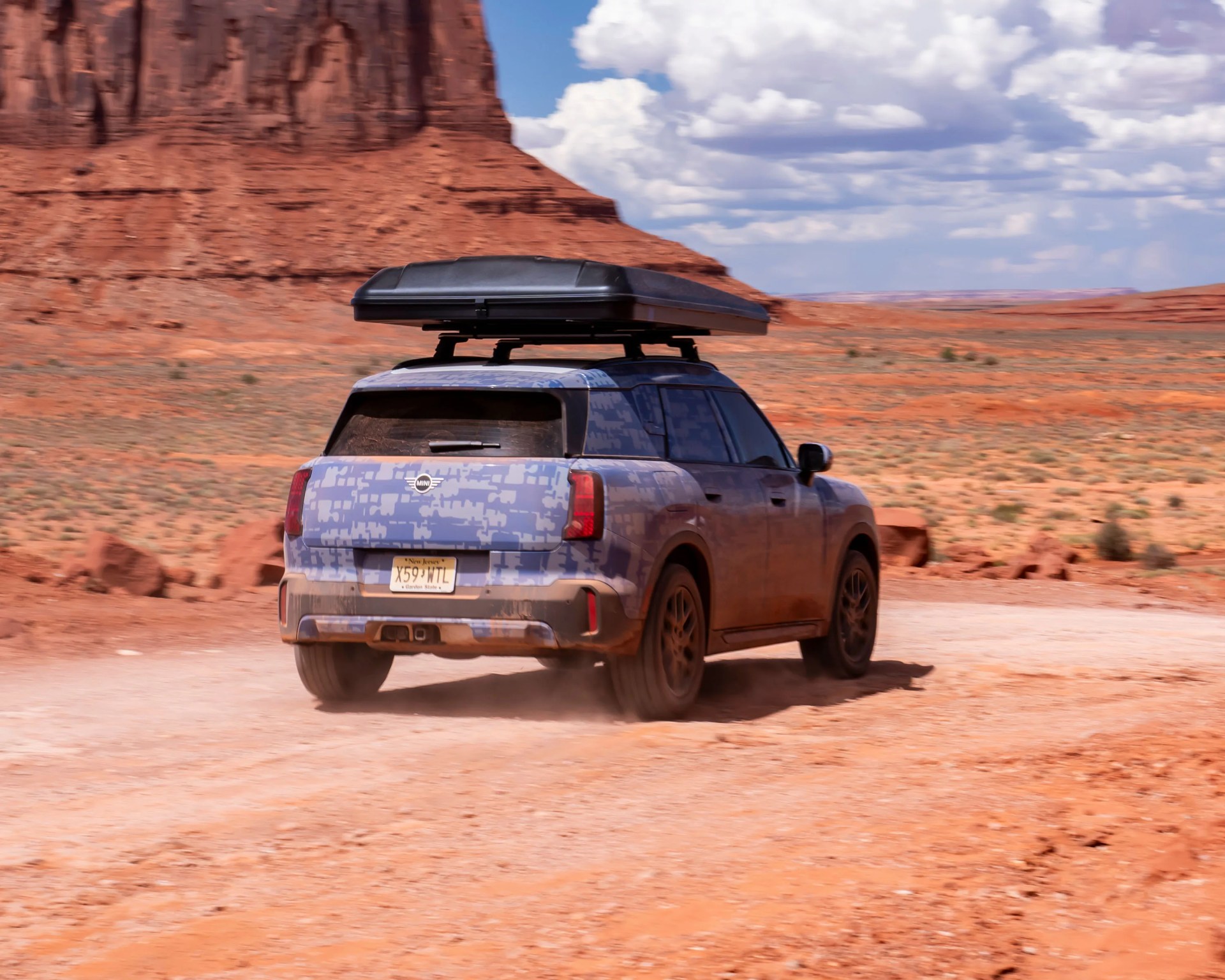 Blue camouflage Mini SUV with black roof cargo box driving on a dusty desert road with red rock formations.