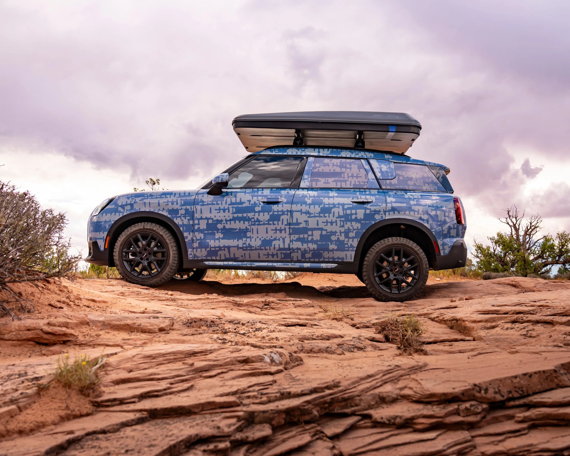 Blue and gray patterned SUV with black roof cargo box on rocky terrain under cloudy sky.