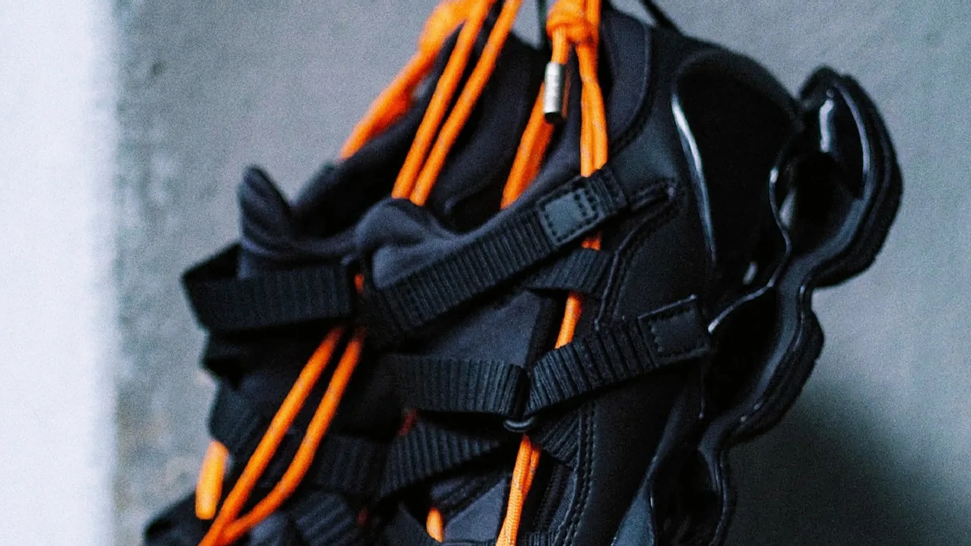 Black sneaker with bright orange laces and multiple black straps on a gray surface.