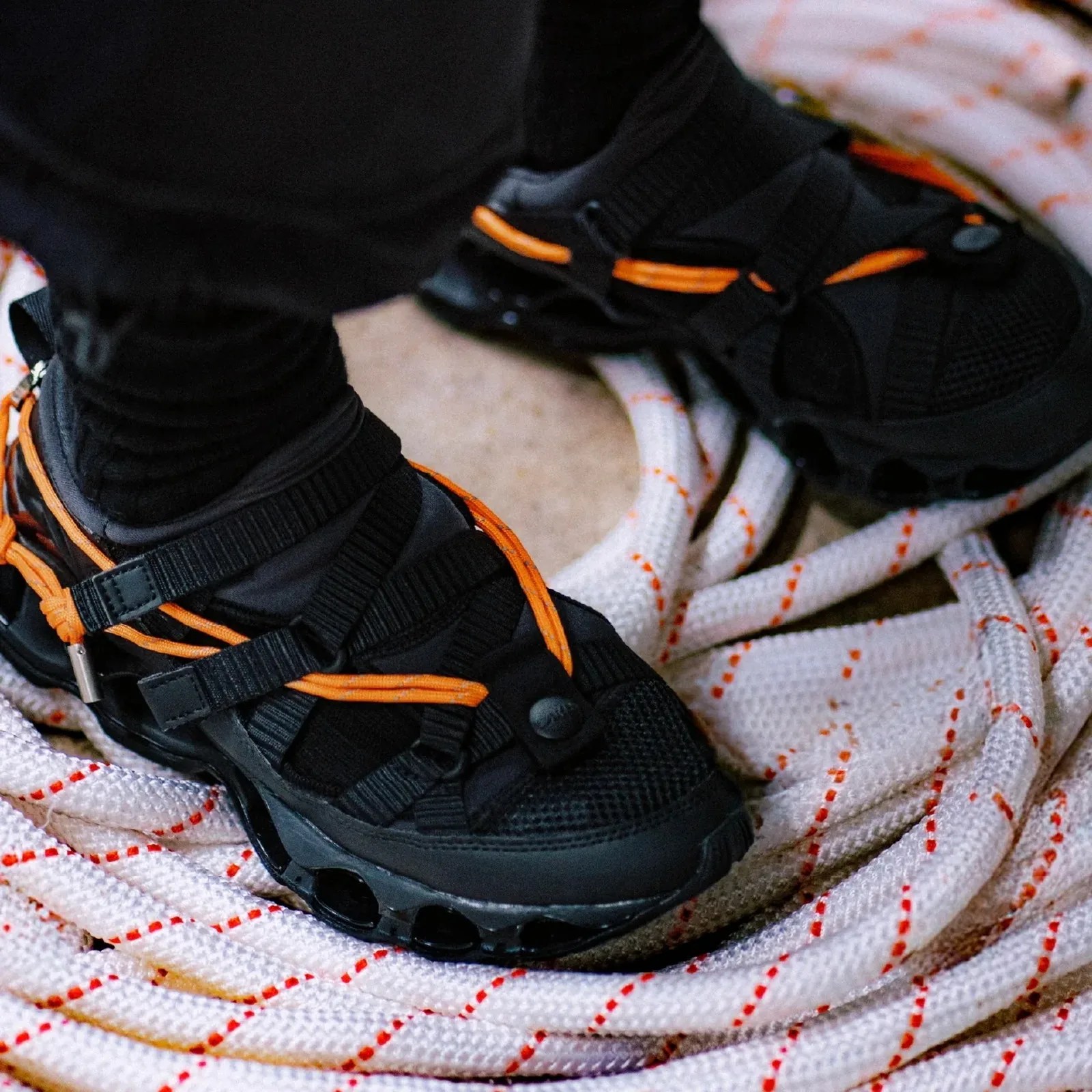 Black and orange outdoor sandals worn on feet standing on coiled white rope with orange markings.