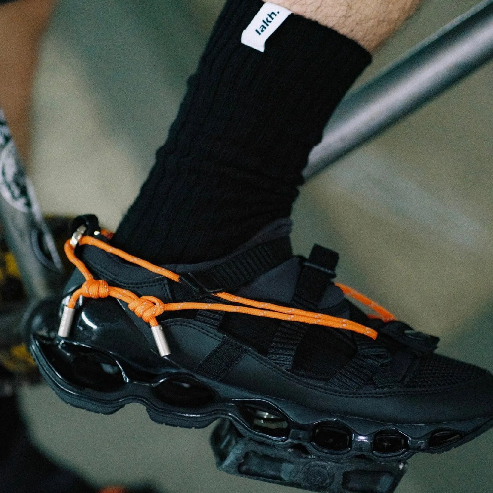 Black sneaker with glossy sole and bright orange laces worn with black ribbed socks.