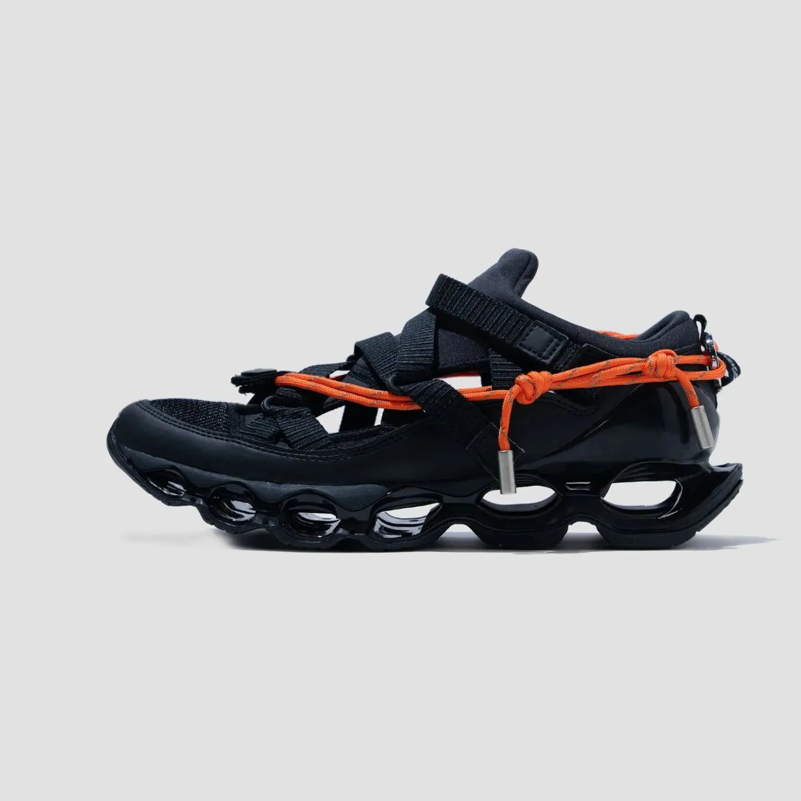 Black sneaker with a hollowed-out sole and orange rope laces on a gray background.