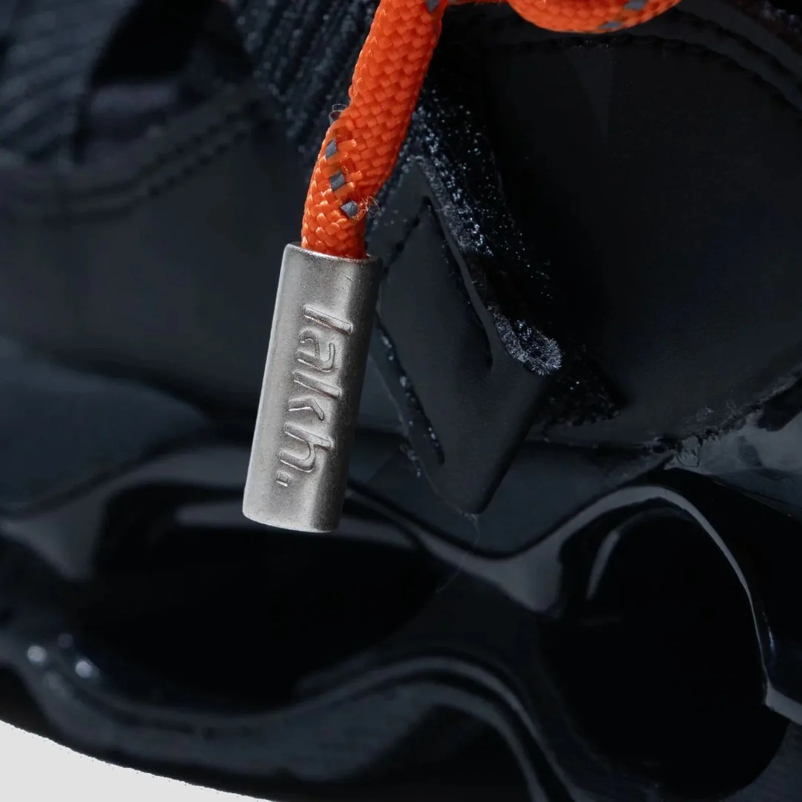 Close-up of an orange shoelace with a silver metal aglet engraved with "larkh" on a glossy black shoe.