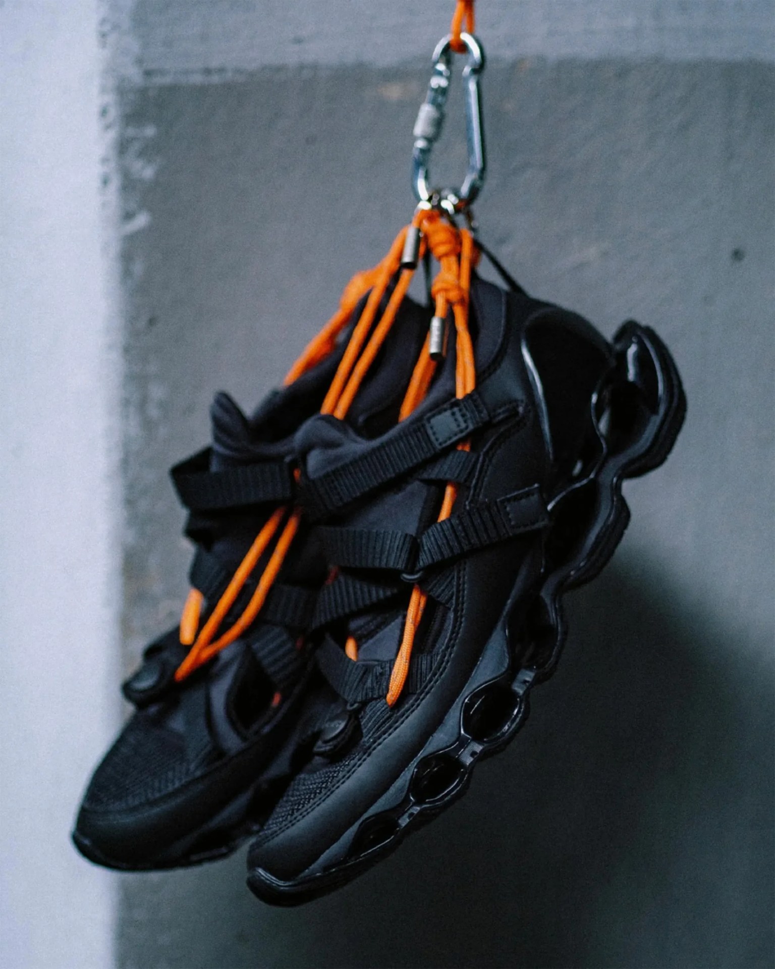 Black athletic shoes with orange laces hanging from a silver carabiner against a gray wall.
