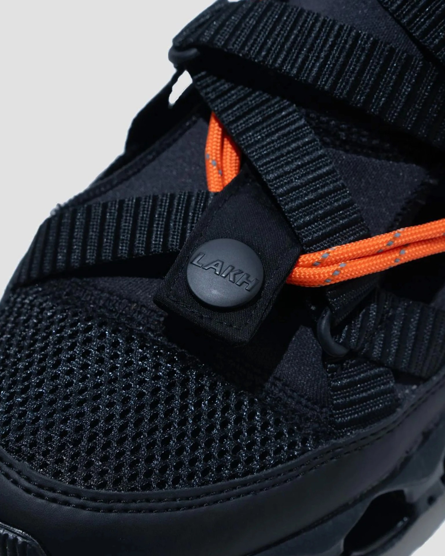 Black mesh sneaker with black straps, orange laces, and a black button labeled "LAKH.