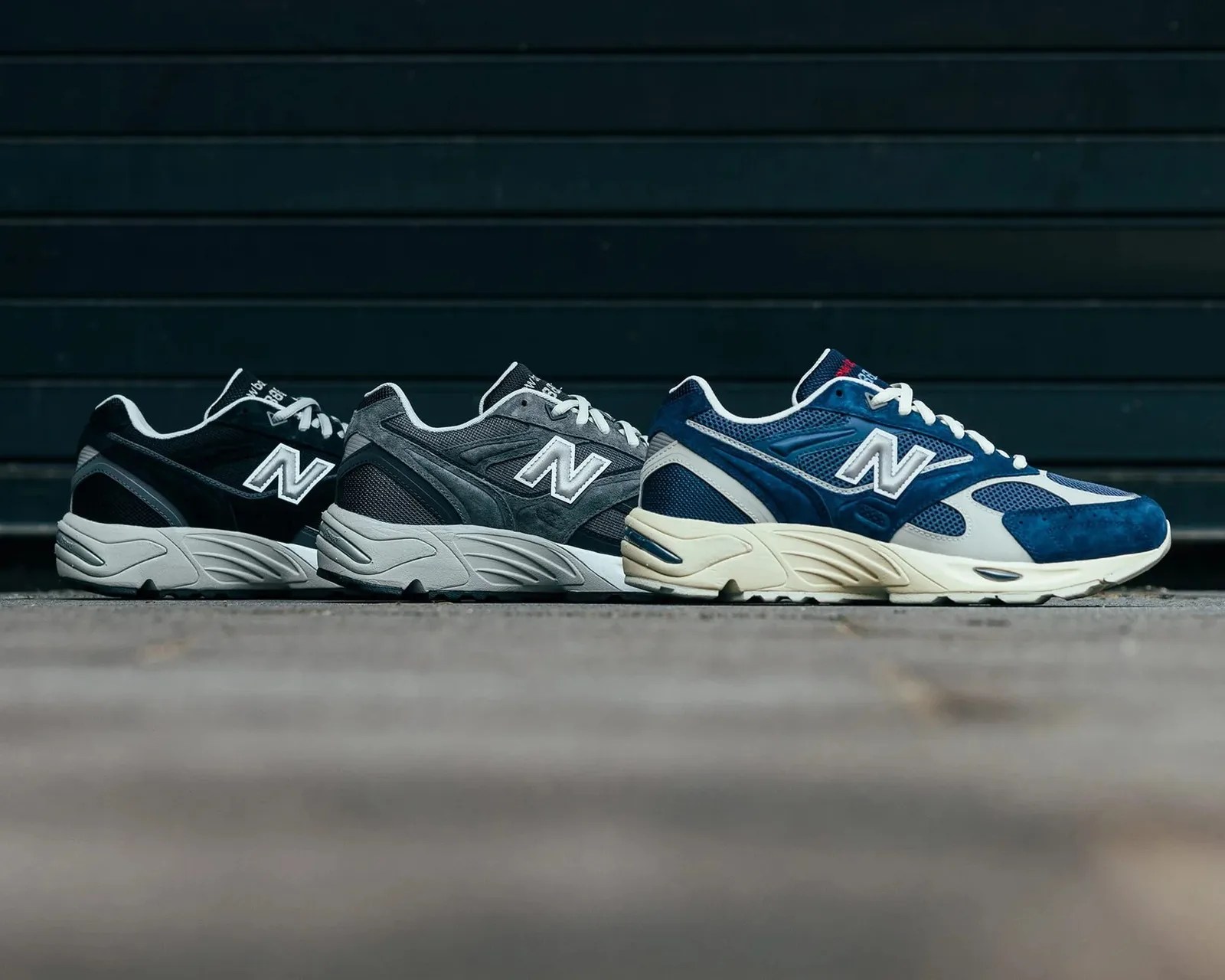 Three pairs of New Balance sneakers in black, gray, and blue with white midsoles lined up on a concrete surface.