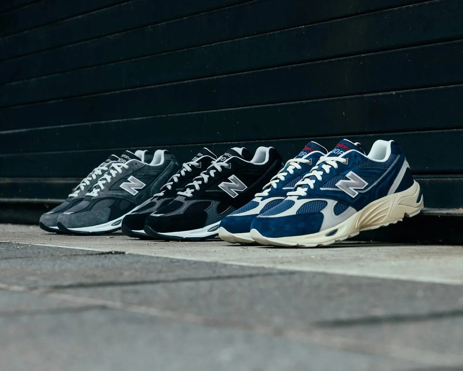 Four pairs of New Balance sneakers in gray, black, and blue colors lined up on a concrete floor against a dark wall.